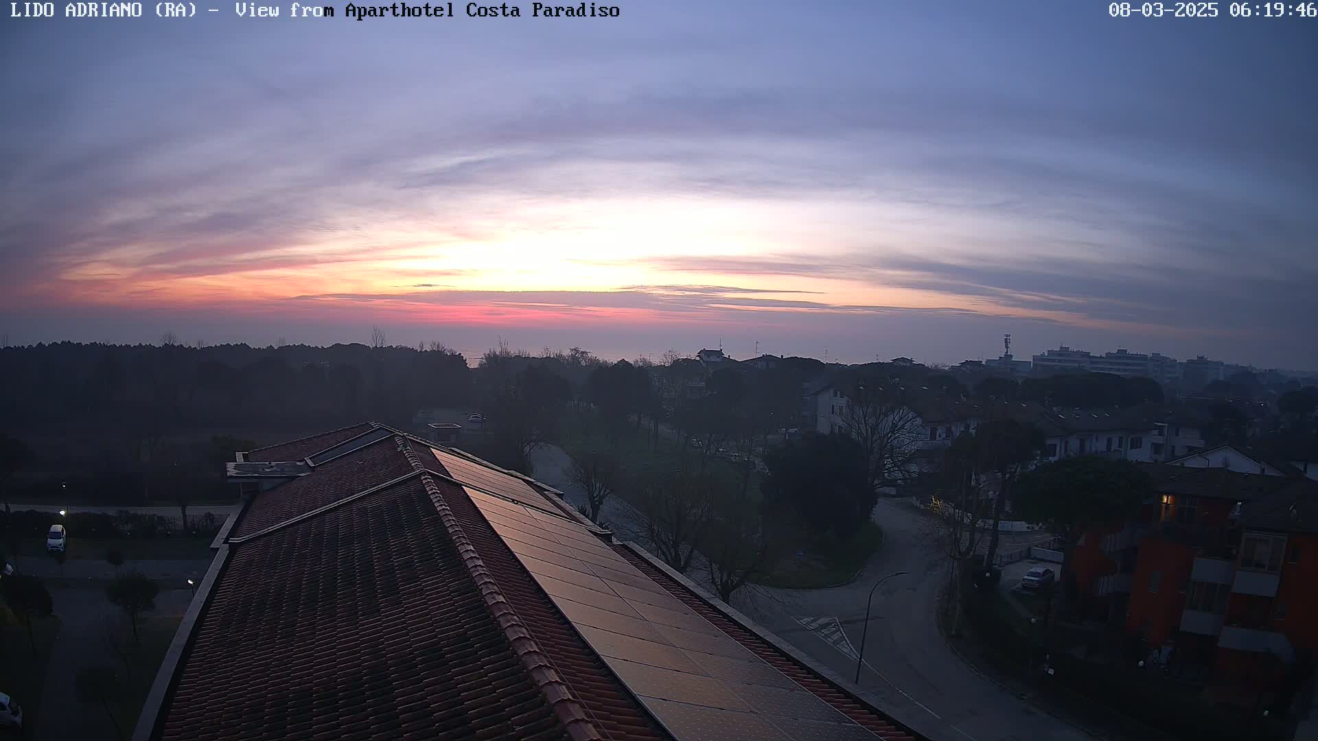 A sunrise with pink and purple hues casts a hazy glow over a residential area viewed from a rooftop with solar panels.