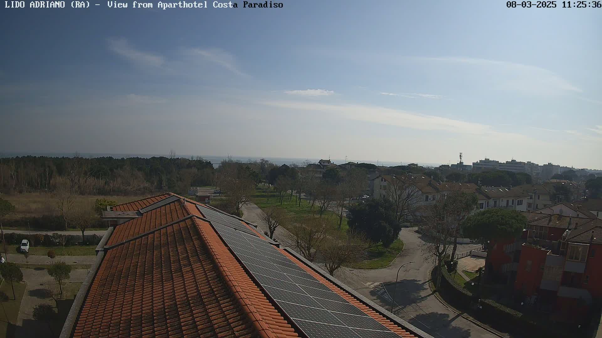A mostly sunny day reveals a coastal town with residential buildings, a park, and a tiled rooftop with solar panels, overlooking a calm sea in the distance.
