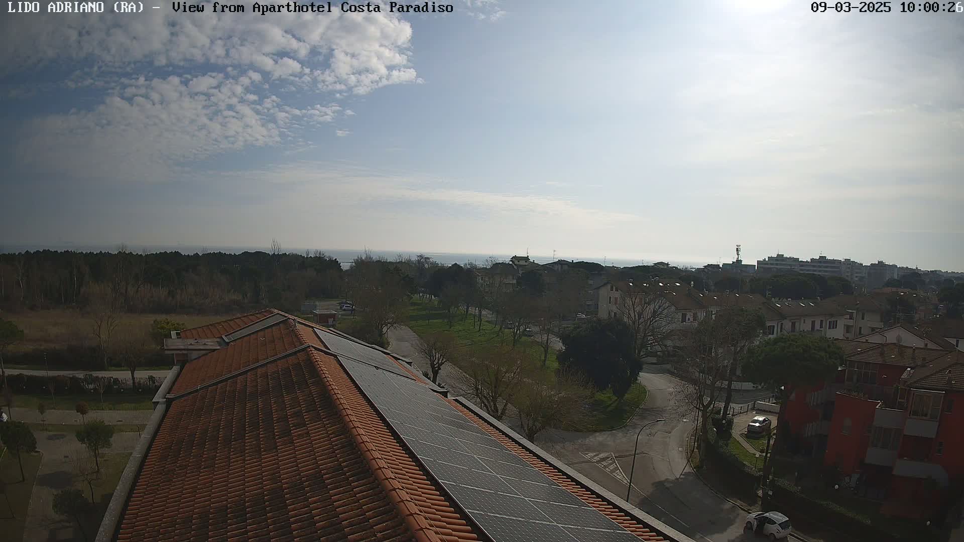 A mostly sunny day reveals a view of a residential area with a sea in the distance, seen from a rooftop with solar panels.
