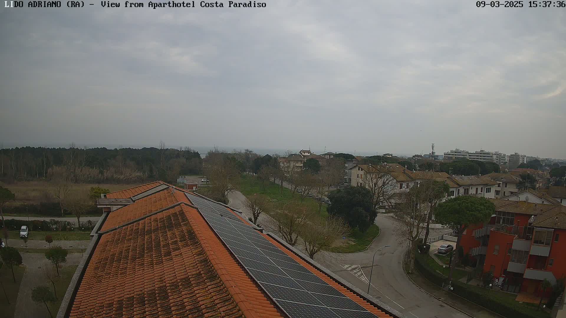 A high-angle view on an overcast day shows a residential area with a tiled roof and solar panels, a street, and trees, overlooking a body of water in the distance.