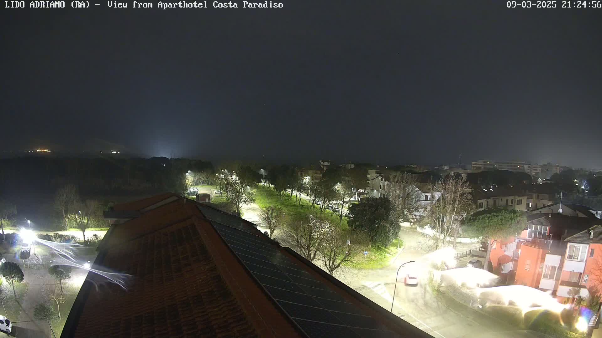 A nighttime, rain-swept view of a street lined with trees and buildings, illuminated by streetlights.