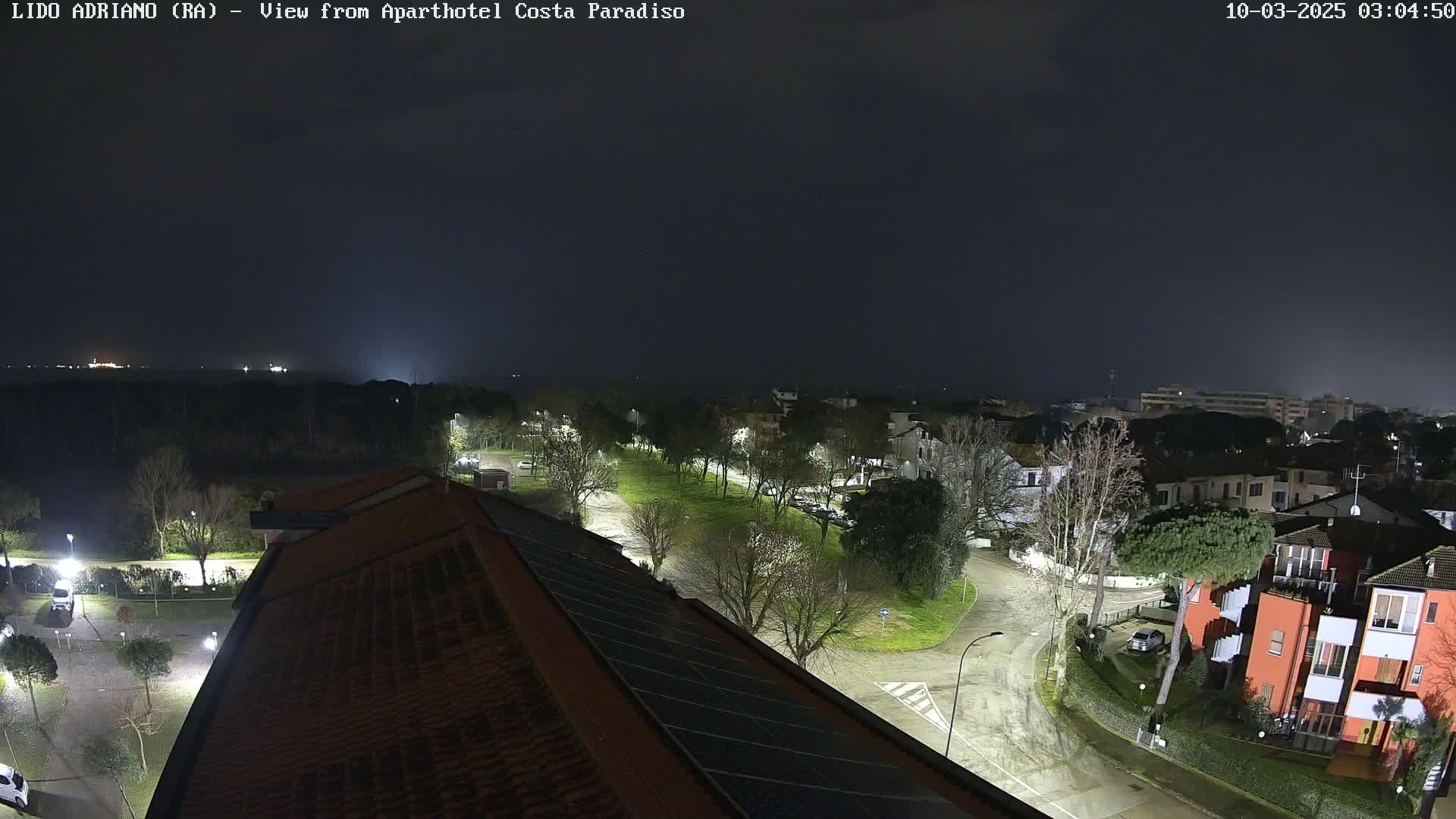 A nighttime view of a residential area with a central park, illuminated streets, and a few cars, under overcast skies.