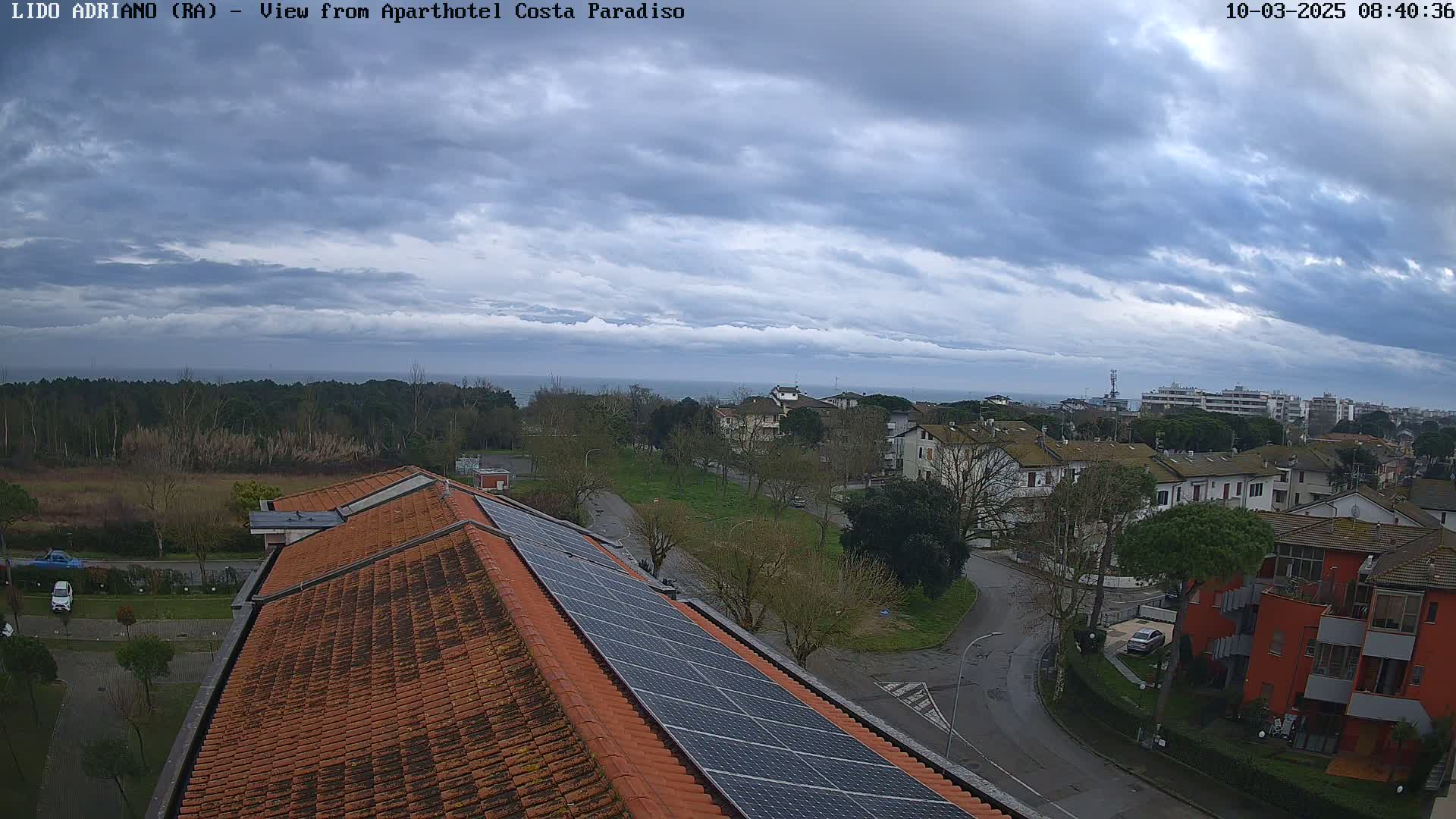 A view from a rooftop with solar panels shows a residential area near a calm sea under a cloudy sky.