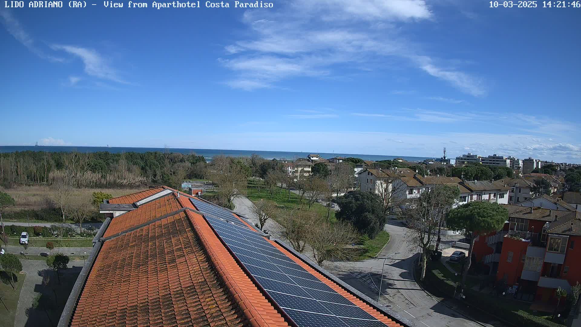 A mostly sunny day shows an aerial view of a building with solar panels on its roof, overlooking a residential area and a calm ocean in the distance.