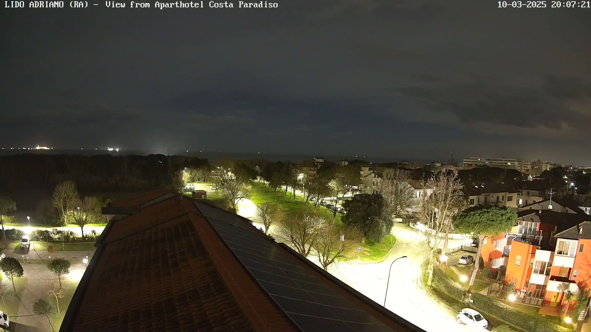 A nighttime aerial view of a residential area near a coast under a partly cloudy sky, showing illuminated streets and buildings.