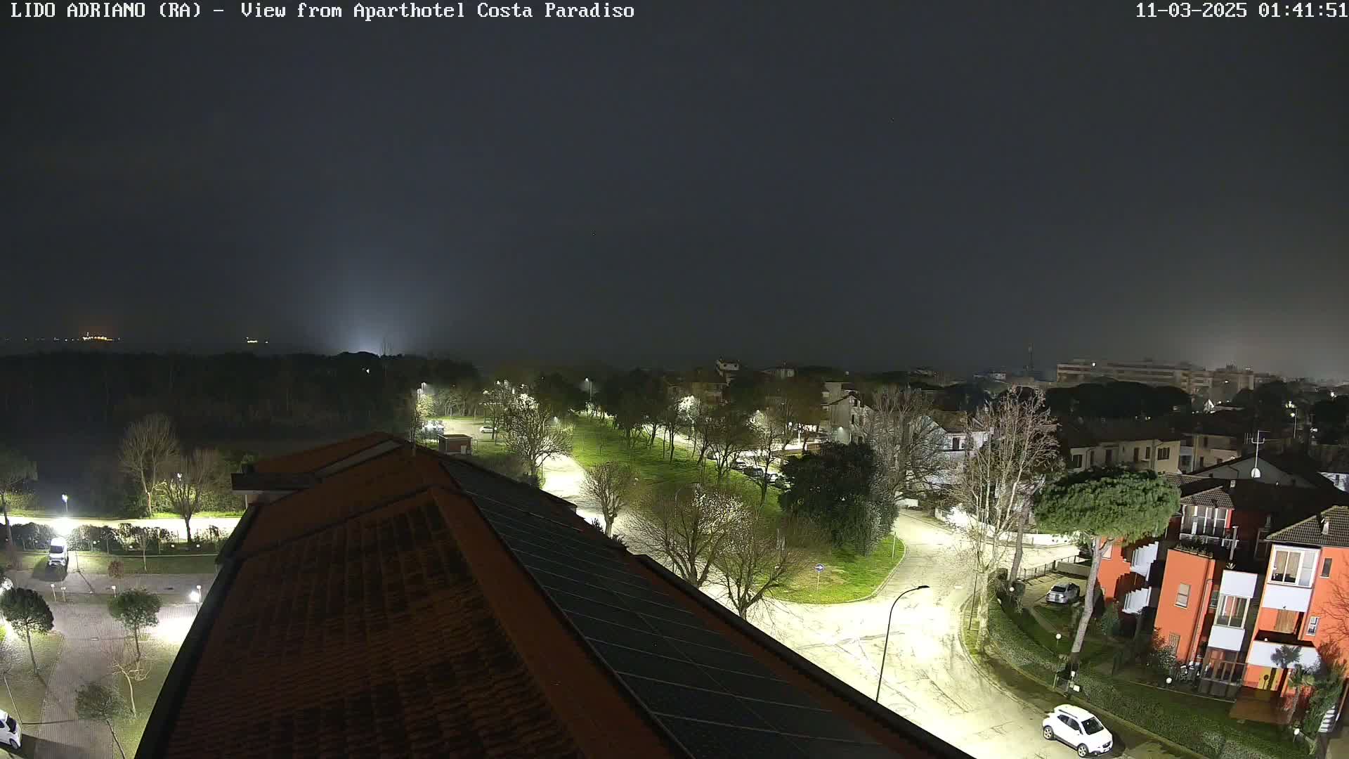 A nighttime view of a residential area with a few cars parked on lightly snow-covered streets under a dark, cloudy sky.