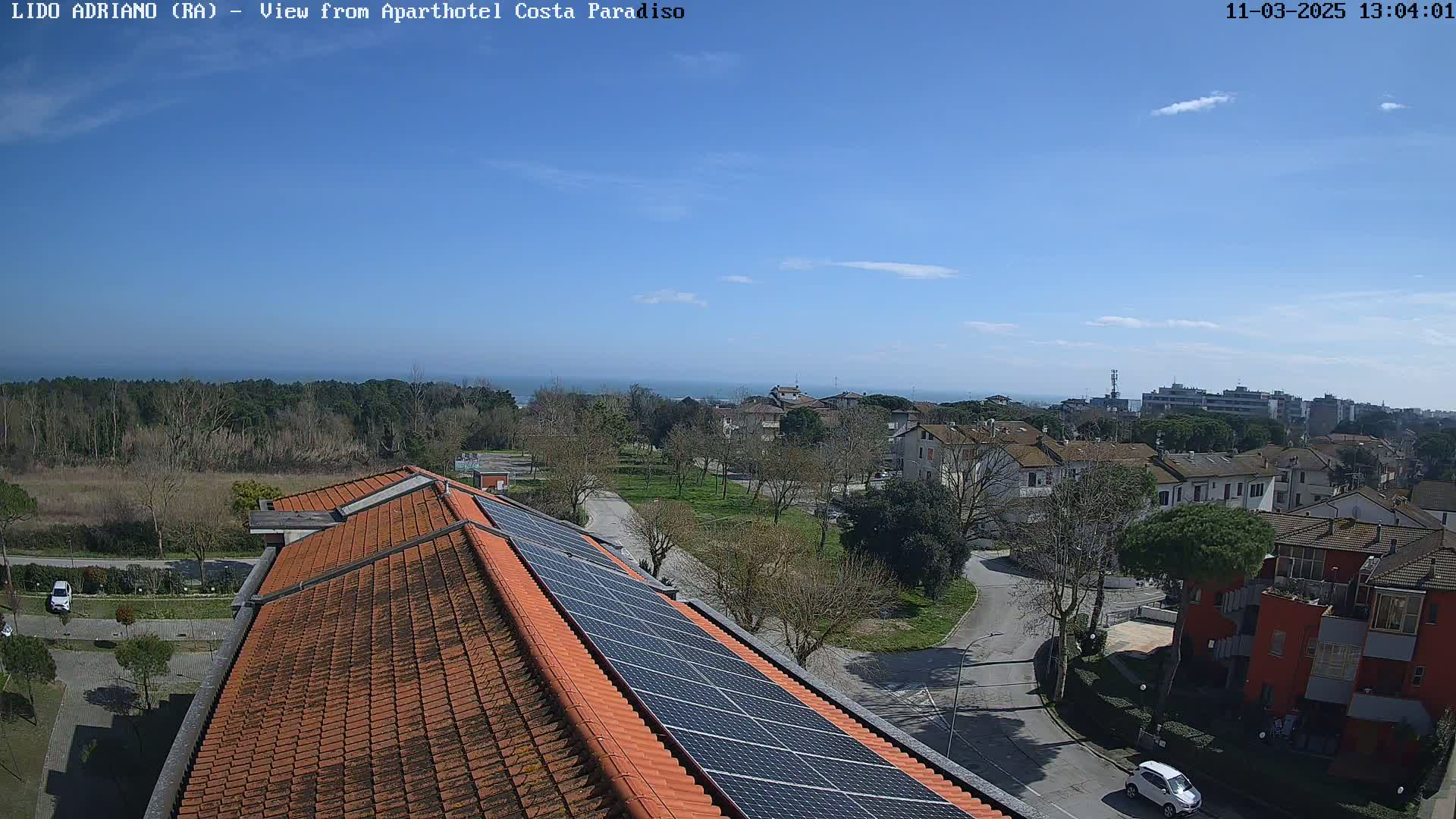 A high-angle view shows a building with a red tile roof and solar panels, overlooking a residential area with a park and a distant ocean under a clear blue sky.
