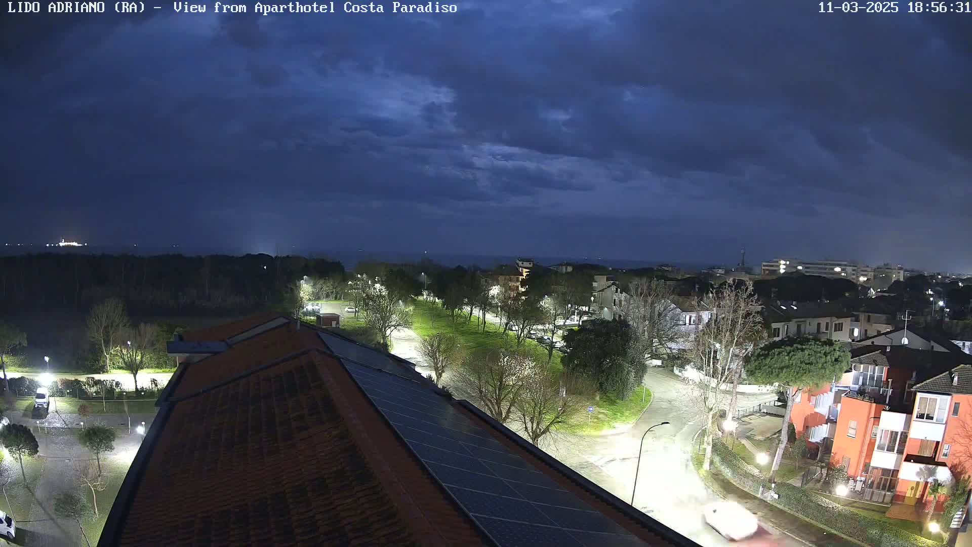 A nighttime view of a coastal town under a dark, cloudy sky shows a residential area with lit streets and buildings, and a dark ocean in the distance.