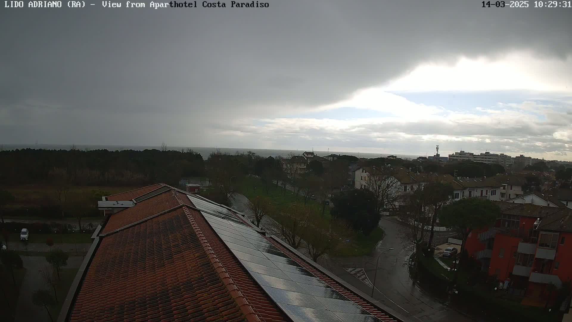 A view of a coastal town on a cloudy, rainy day, seen from a rooftop with solar panels, showing a calm sea in the distance.