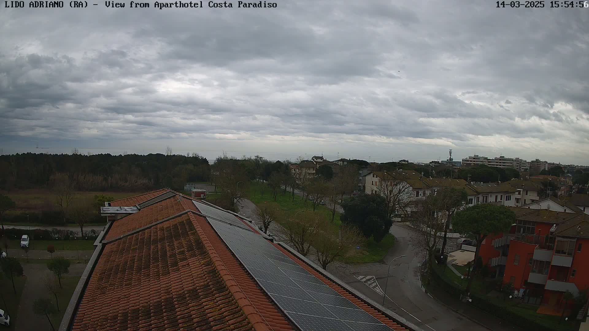 A view of a residential area with a tiled roof and solar panels on top, overlooking a calm sea under a cloudy sky.