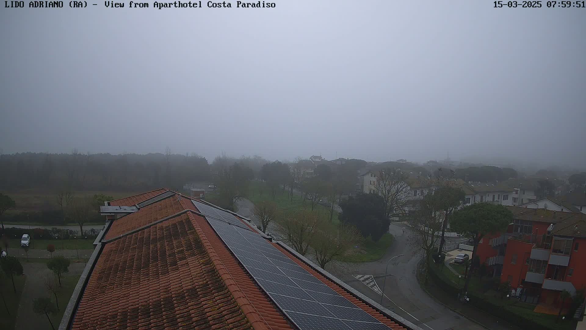 A foggy view of a residential area with a red building and solar panels on a tiled roof in the foreground.
