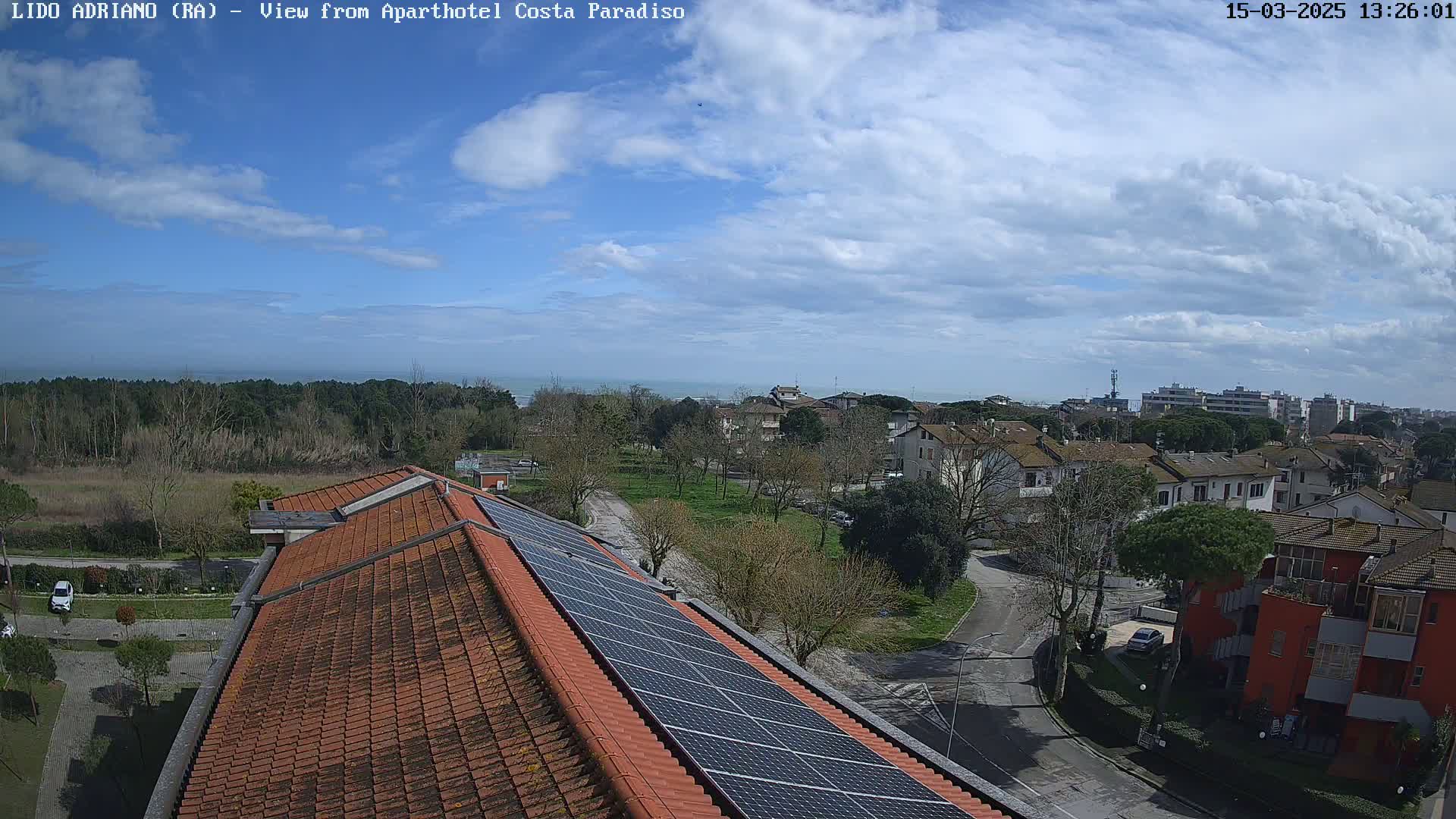 A partly cloudy day reveals a coastal town with residential buildings, a tree-lined park, and a rooftop with solar panels, overlooking a calm sea in the distance.