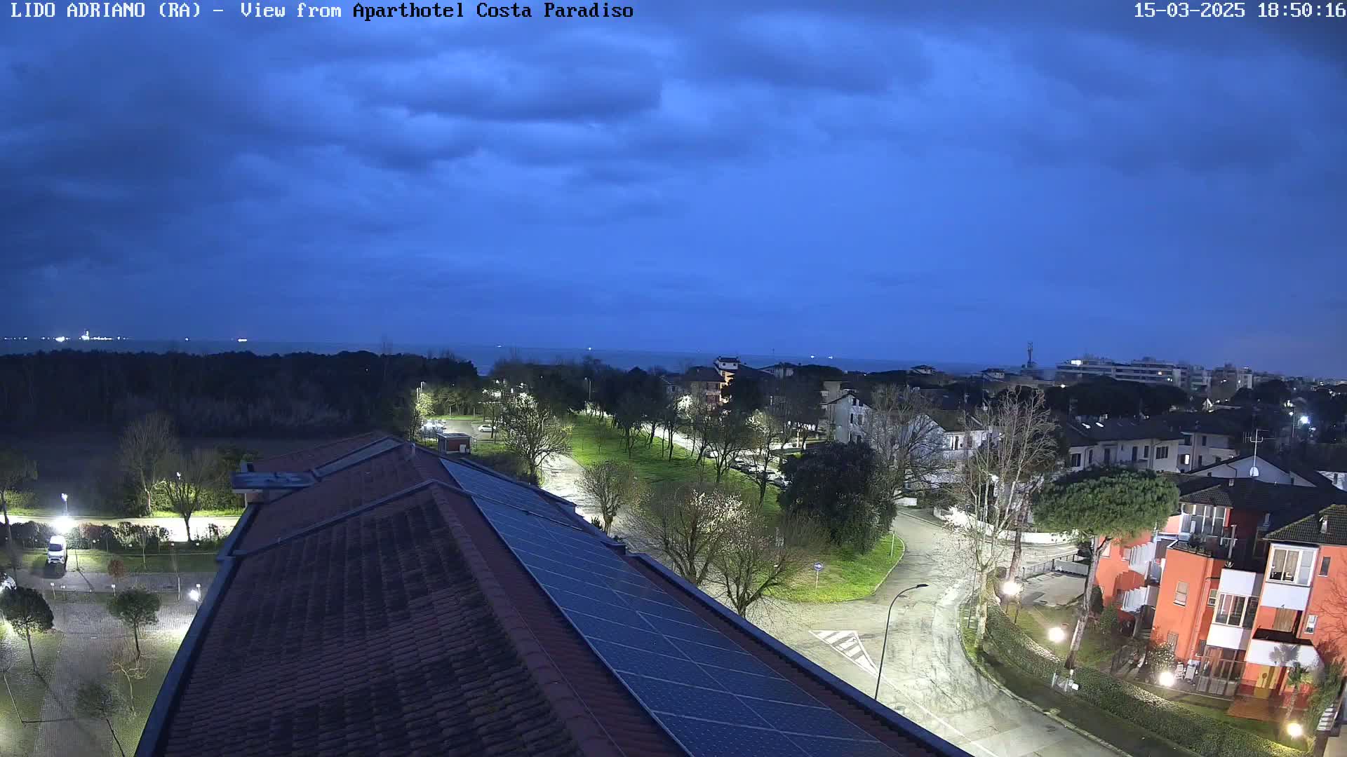 A nighttime view of a residential area near a coast under a mostly clear, dark blue twilight sky shows a street curving through the neighborhood, with houses and trees illuminated by streetlights, and a section of a building's roof with solar panels in the foreground.