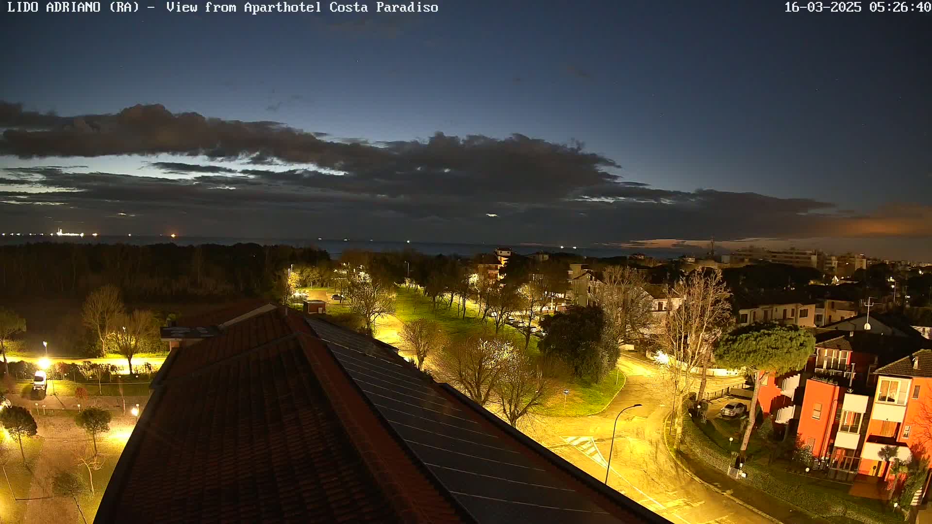 A nighttime view of a residential area near a calm sea under a partly cloudy sky.