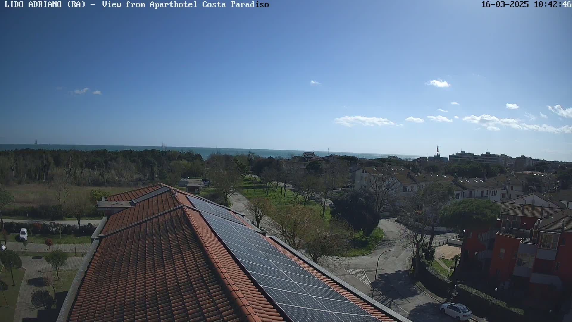 A mostly sunny day overlooks a coastal town with a view of the sea from a rooftop with solar panels.