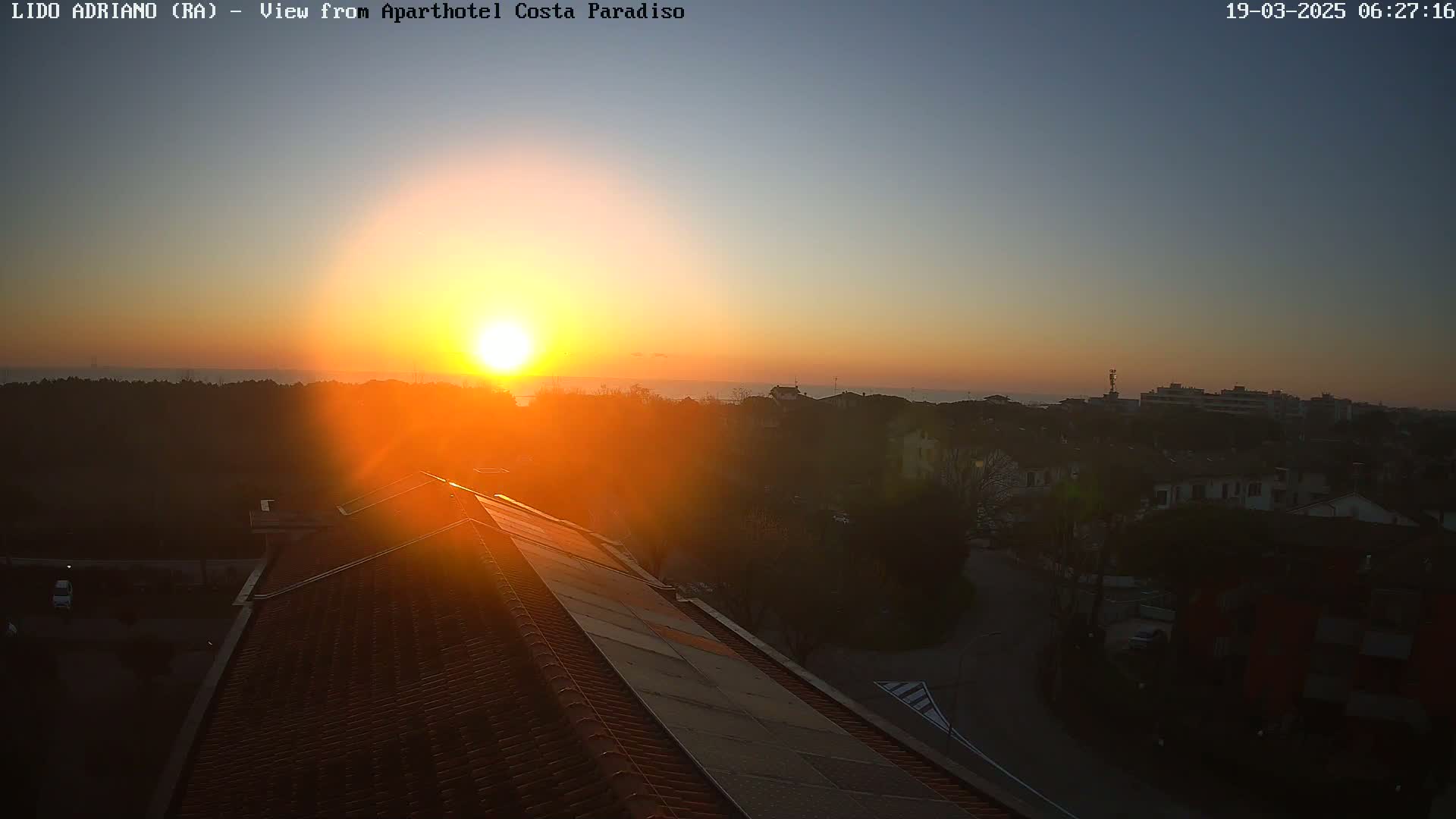 A vibrant sunrise casts an orange glow over a coastal town, seen from a rooftop with a tiled roof.
