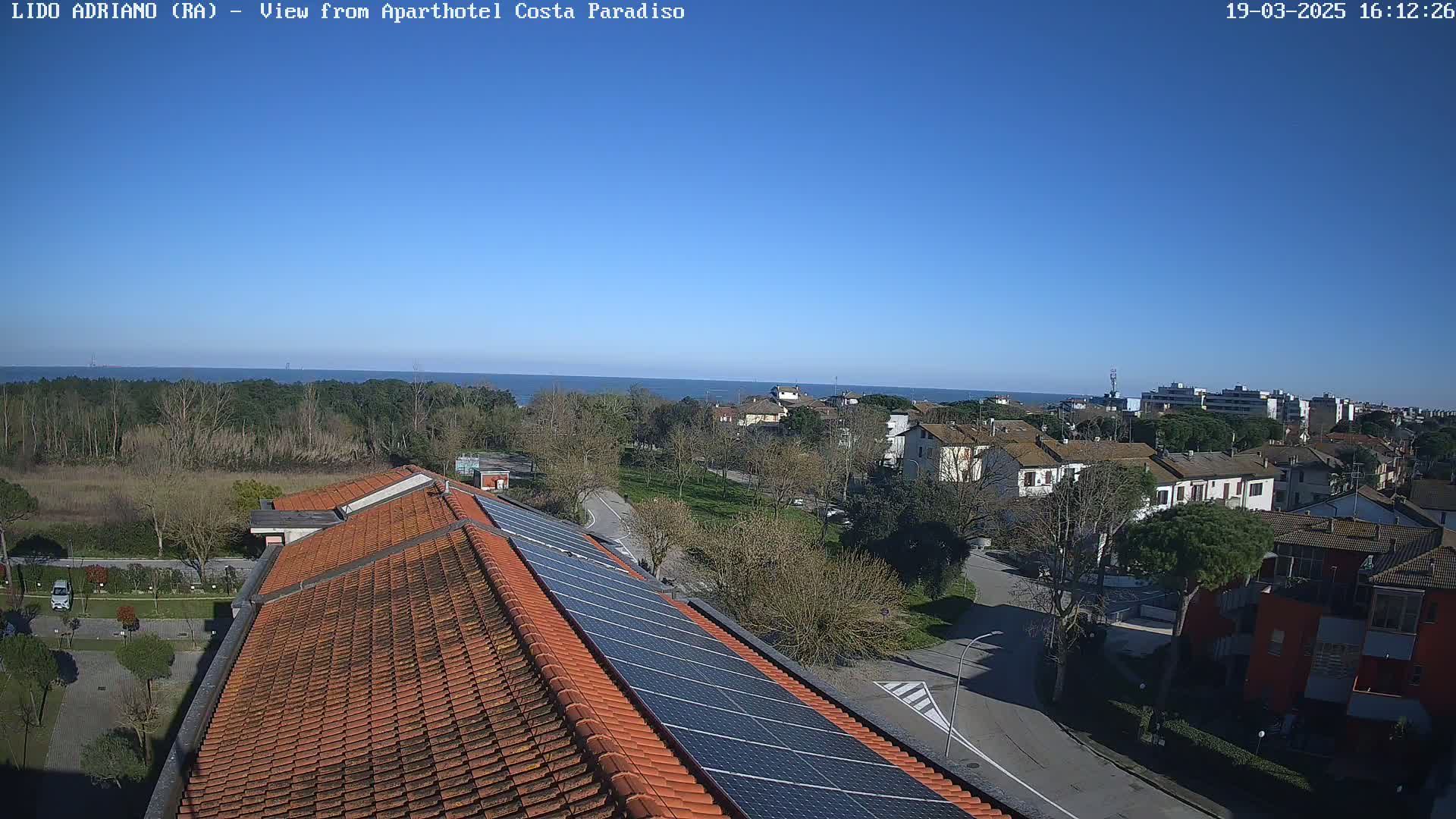 A clear, sunny day overlooking a residential area with a coastline in the distance, showing a building roof with solar panels in the foreground.