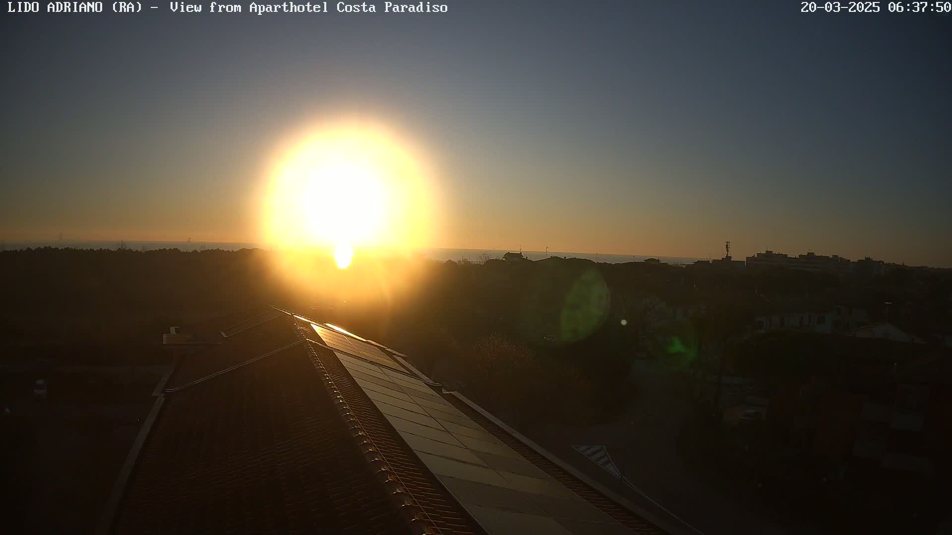 A bright sunrise illuminates a tiled roof with solar panels, overlooking a town and calm body of water in the distance.