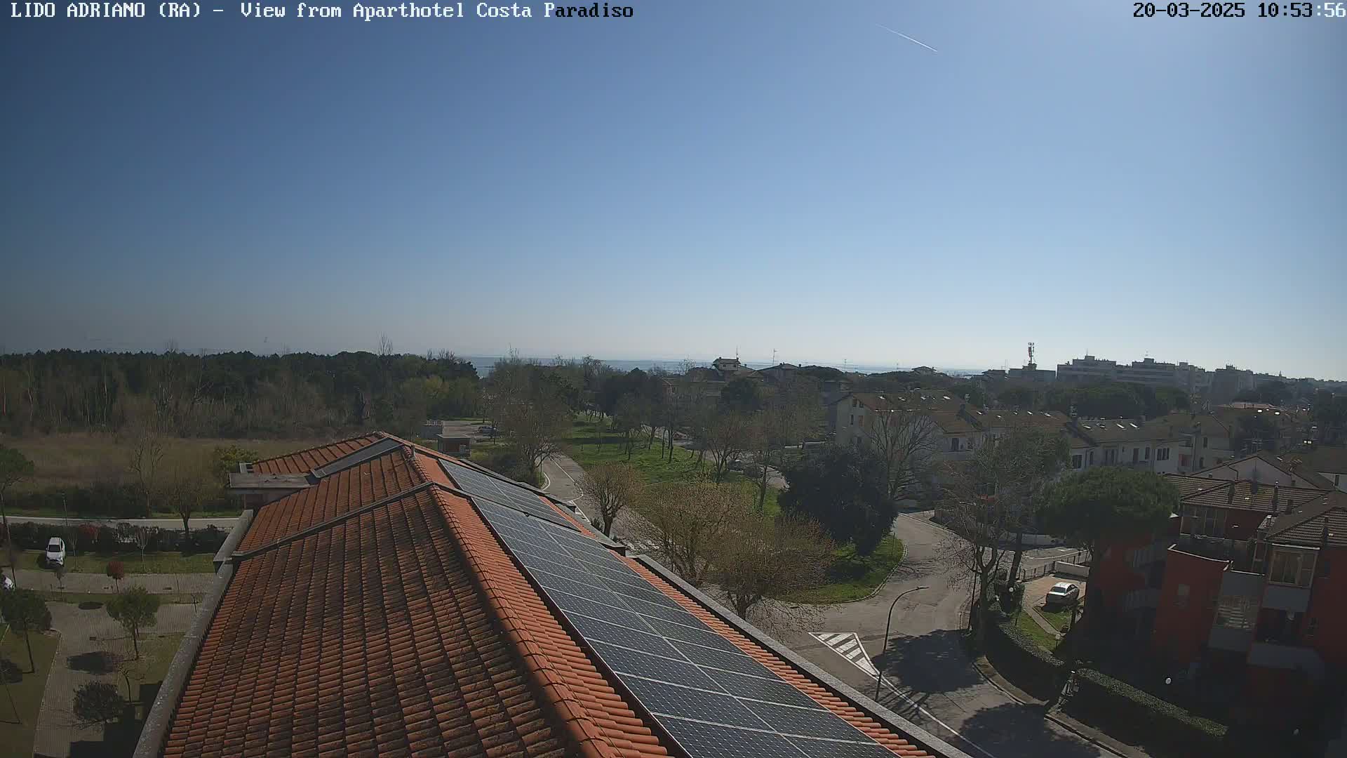 A clear, sunny day reveals a view of a residential area with a coastline in the distance, seen from a rooftop with solar panels.