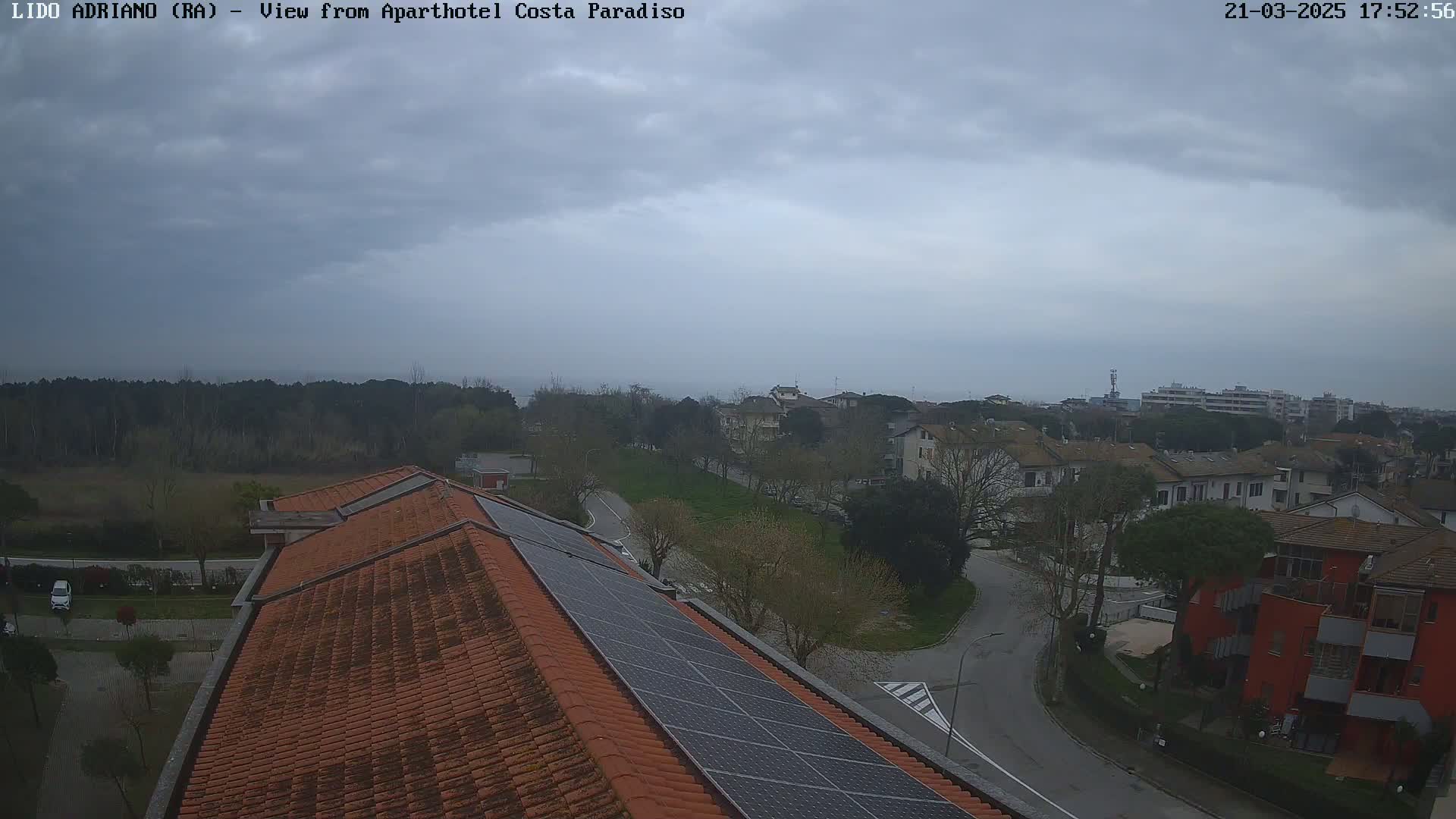 An overcast sky overlooks a residential area with a coastline visible in the distance, featuring a building with solar panels on its roof.