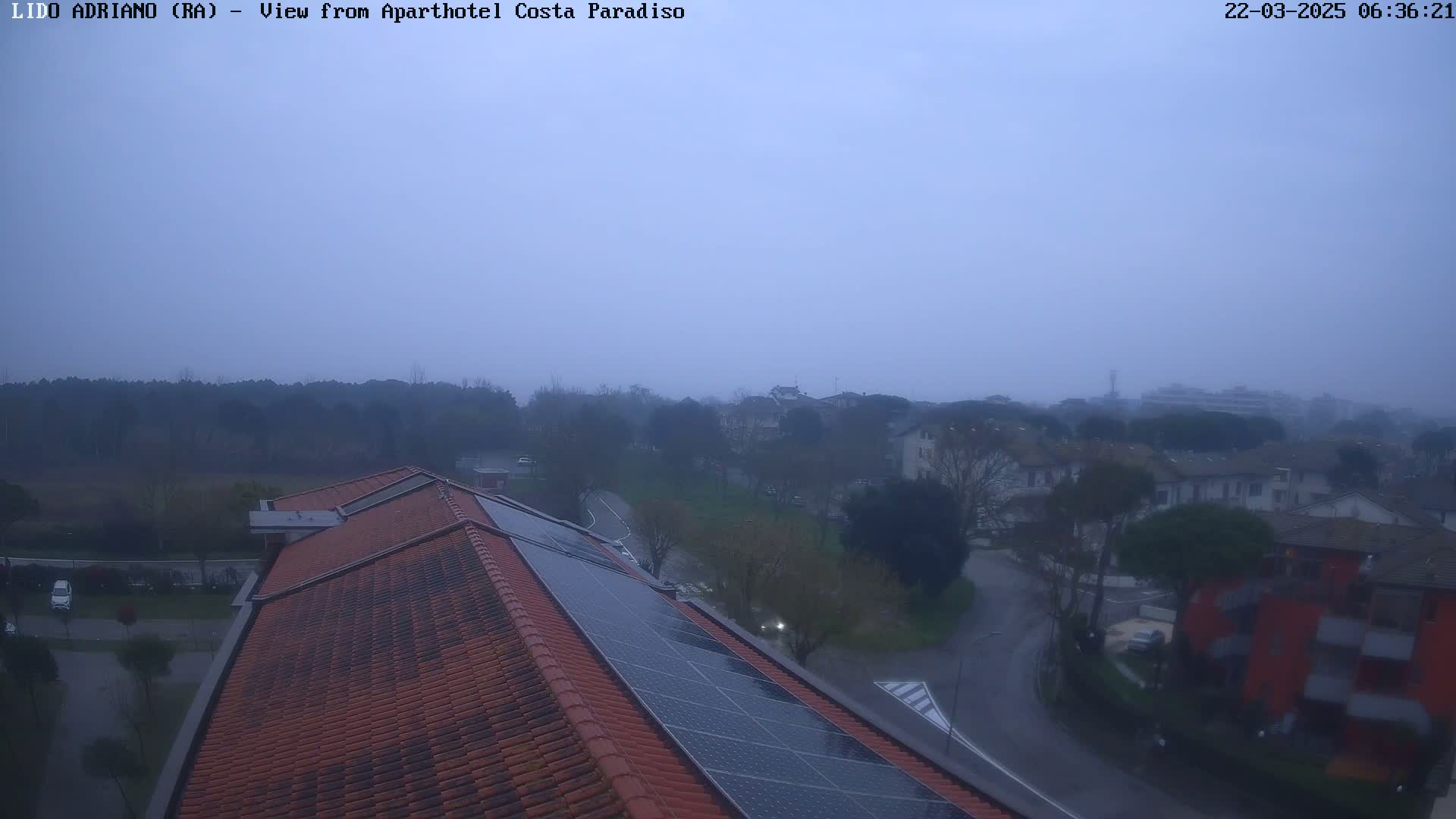 A hazy, overcast view from a rooftop with solar panels shows a residential area with trees and roads.