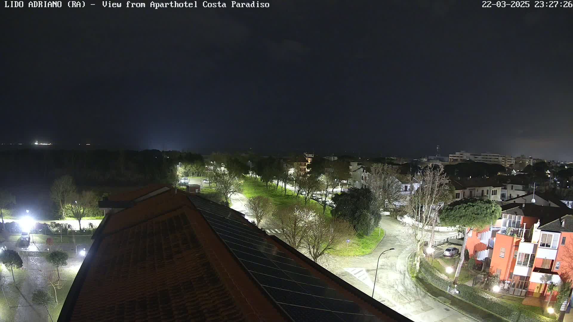 A nighttime, rain-slicked view of a residential area with streetlights illuminating trees, roads, and buildings, extending to a dark body of water in the distance.
