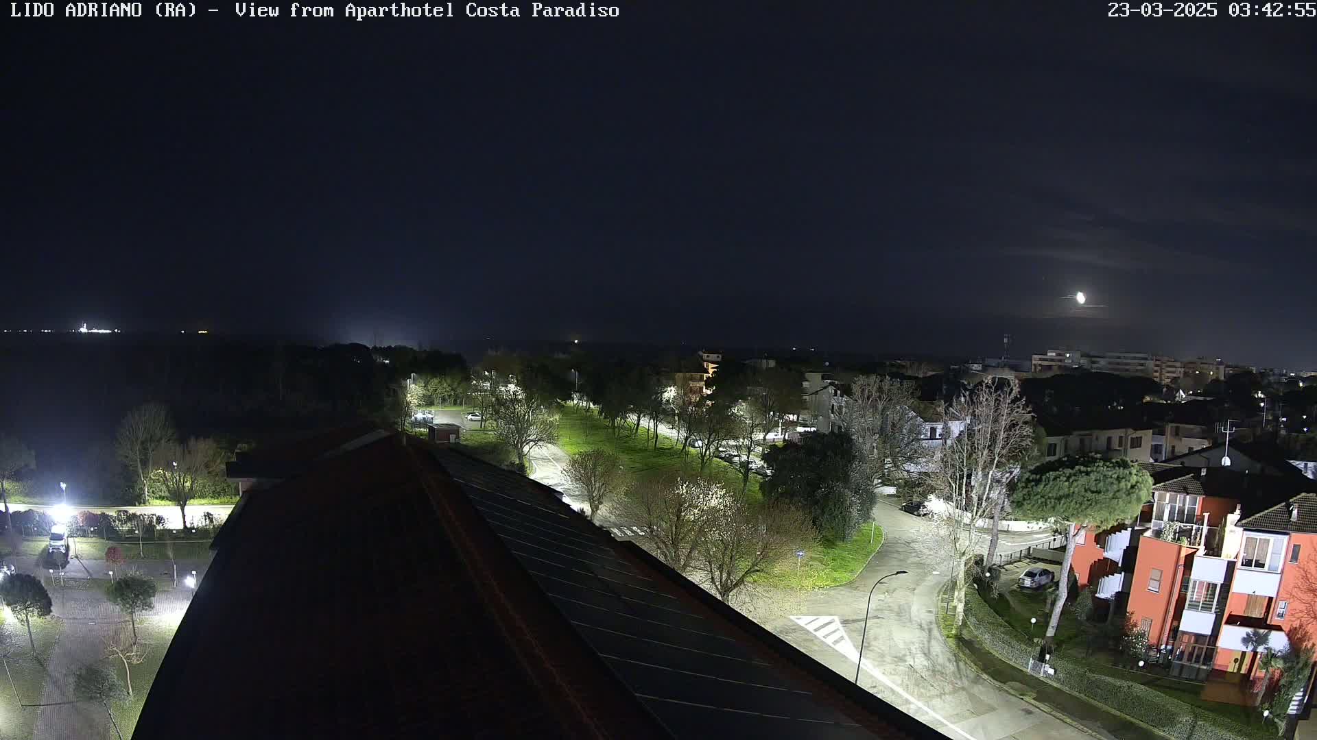 A nighttime, overhead view of a residential area with roads and trees, under a slightly cloudy moonlit sky.