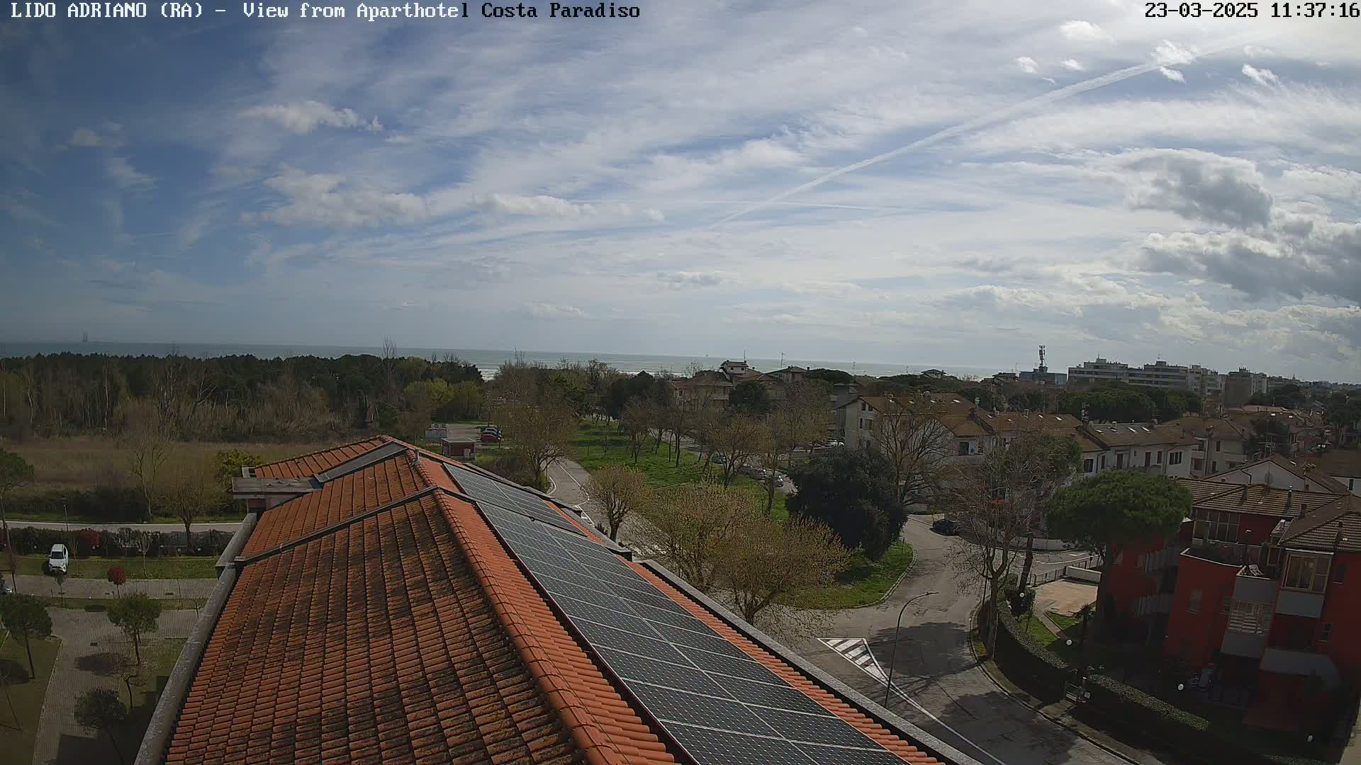 A partly cloudy day reveals a coastal town viewed from a rooftop with solar panels, showing a calm sea in the distance.