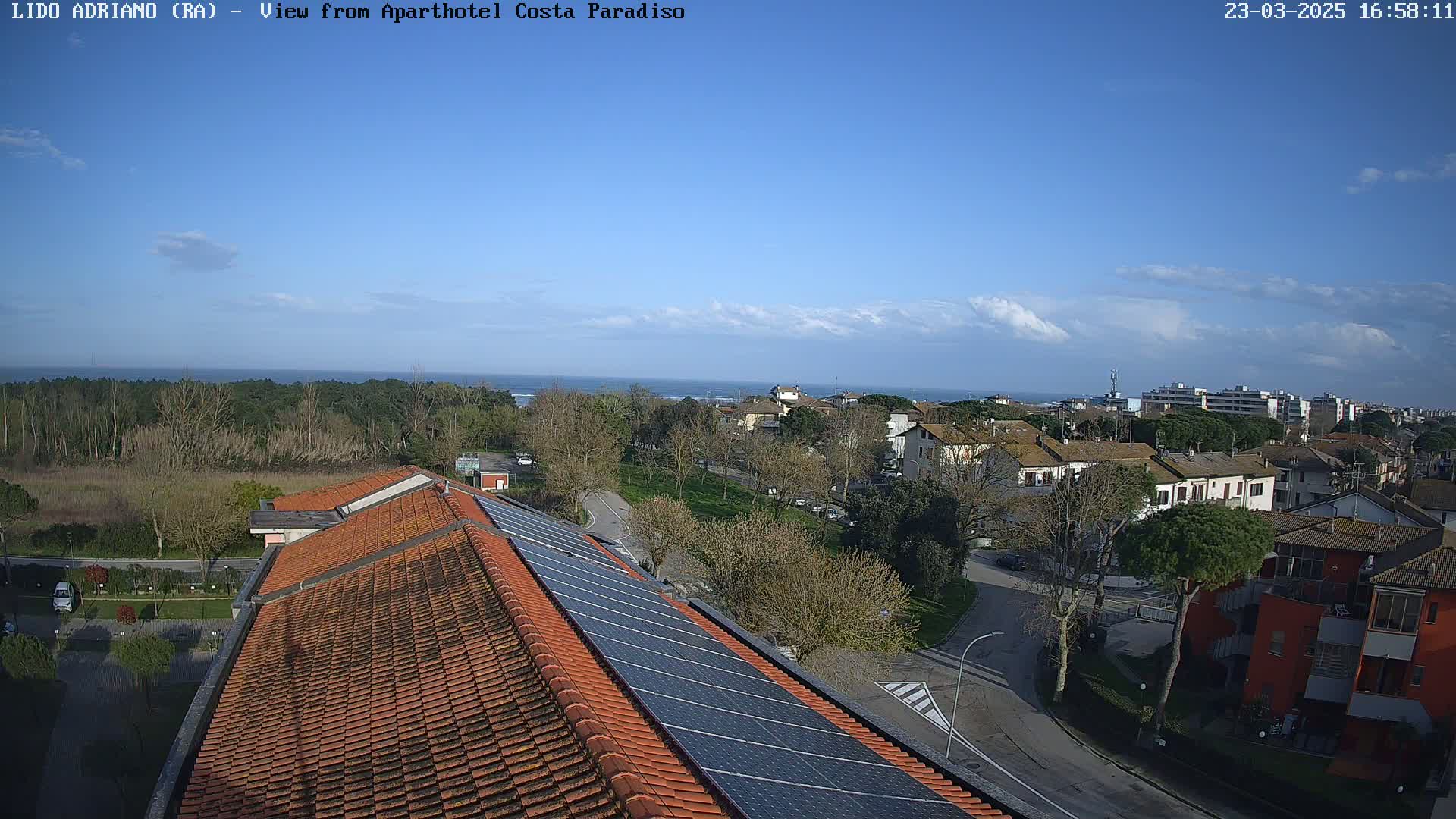 A partly sunny day overlooks a coastal town with residential buildings, trees, and a rooftop featuring solar panels, with a calm ocean visible in the distance.