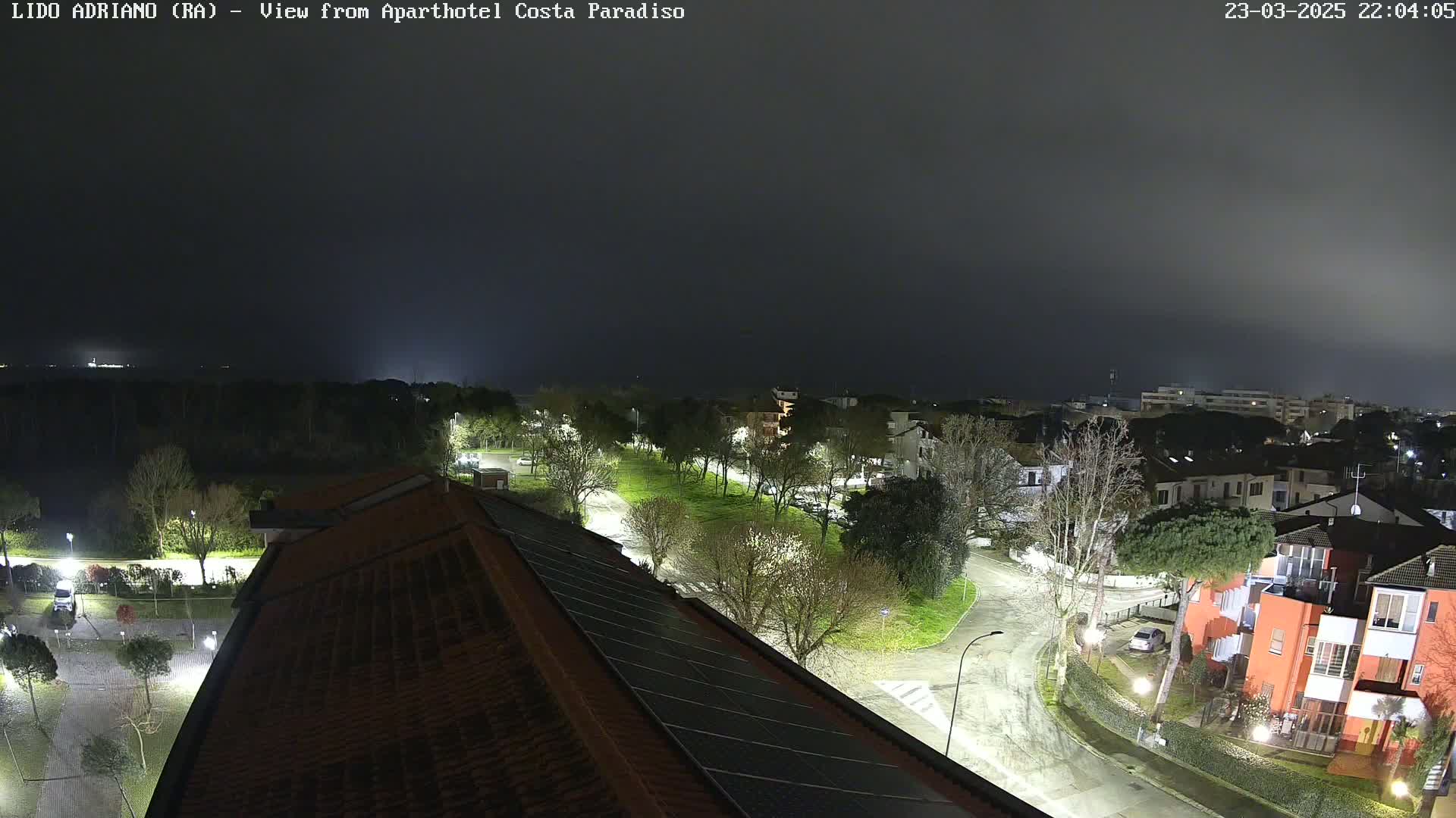A nighttime view of a residential area with a park, illuminated streets, and a building with solar panels on its roof, under a dark, foggy sky.