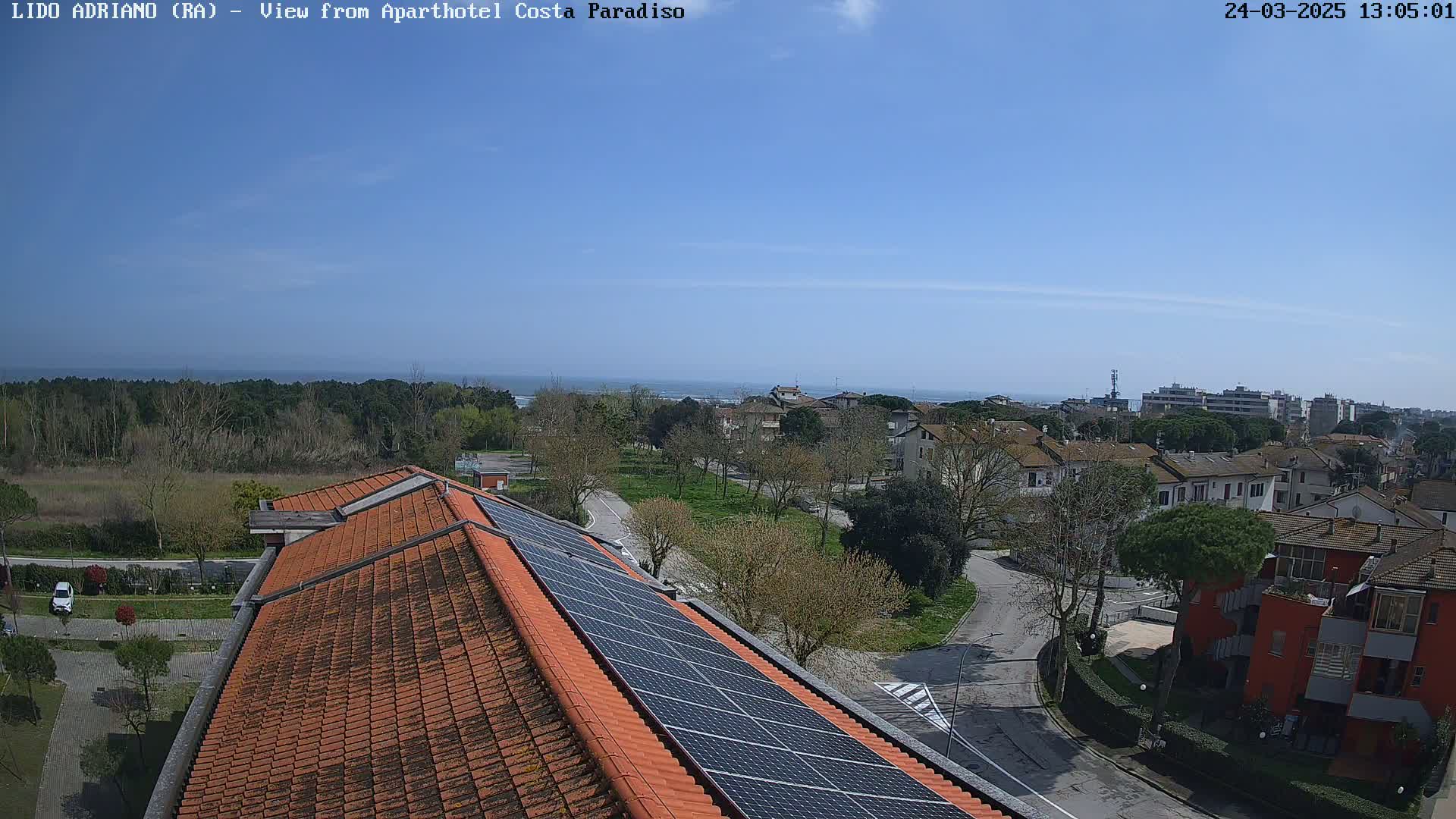 A clear, sunny day overlooks a coastal town with a red-tiled roof and solar panels, showcasing a calm ocean in the distance.
