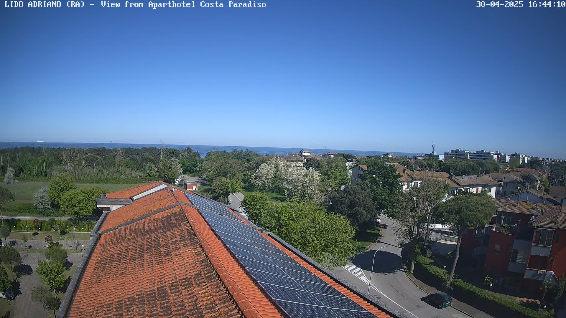 A rooftop with solar panels overlooks a residential area with green trees and a calm blue sea under a clear blue sky.