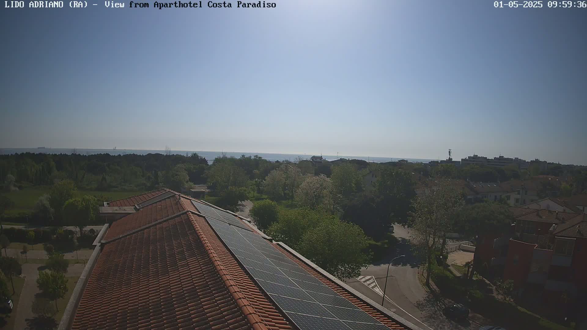 A clear, sunny day overlooks a residential area with a reddish-brown tiled roof and solar panels, extending to a calm ocean in the distance.