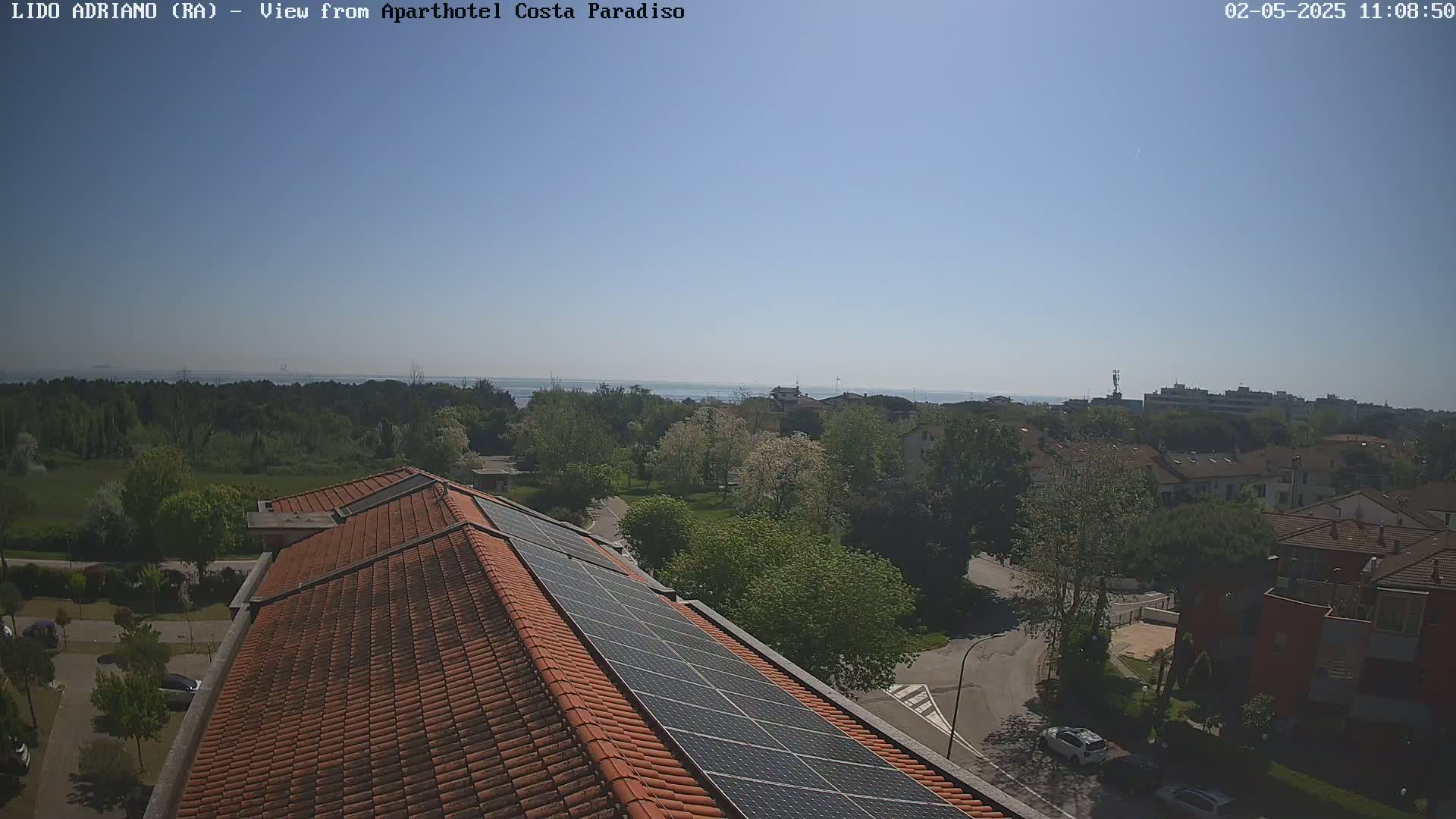 A clear, sunny day overlooks a residential area with a view of the sea in the distance, showing a rooftop with solar panels in the foreground.