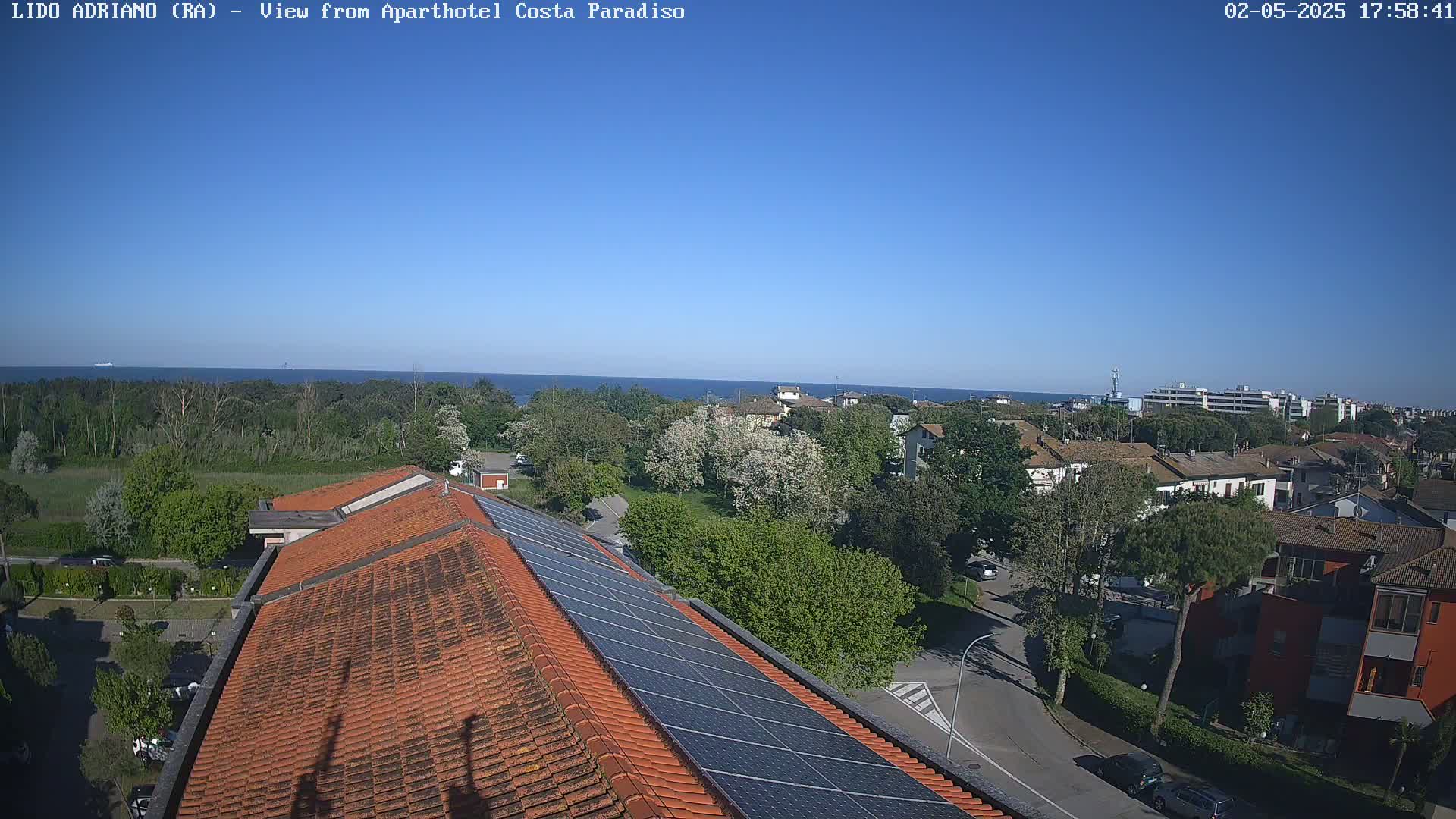 A clear, sunny day reveals a seaside town viewed from a rooftop with solar panels, showing a calm ocean in the distance and lush greenery surrounding buildings.