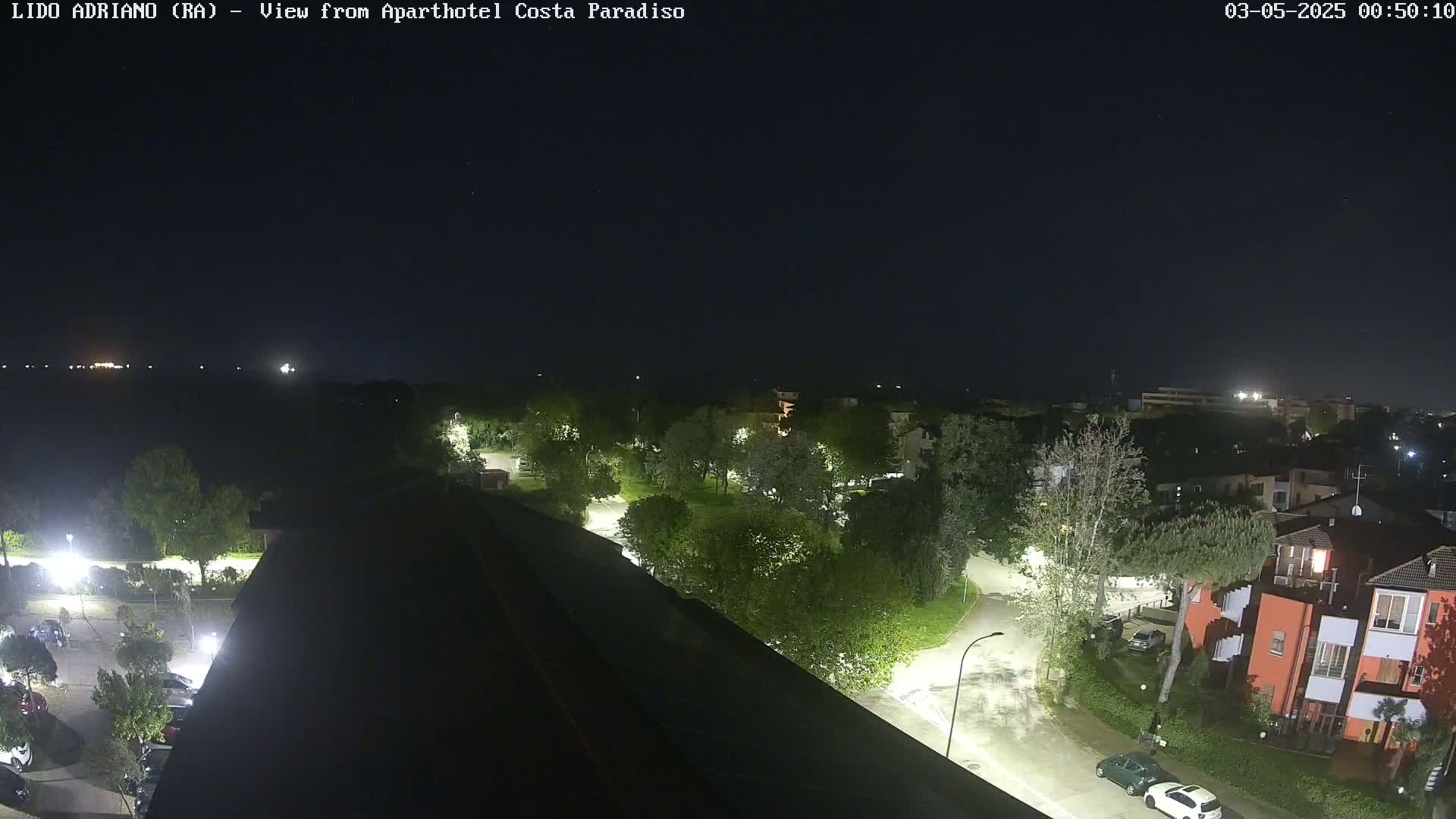 A nighttime view of a residential area with streetlights illuminating trees, roads, and parked cars, under a clear, dark sky.