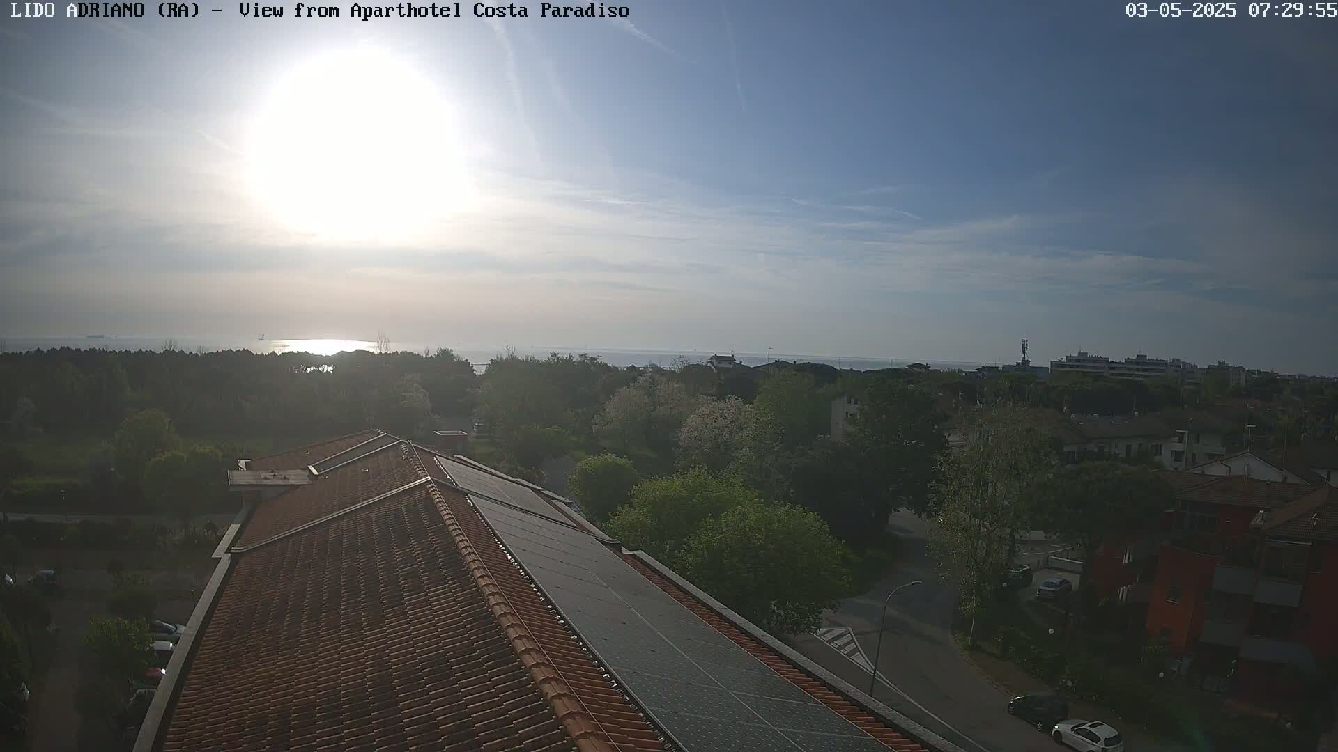 A mostly sunny, slightly hazy day reveals a view of a calm sea beyond a tree line and rooftops dotted with solar panels, overlooking a residential area.