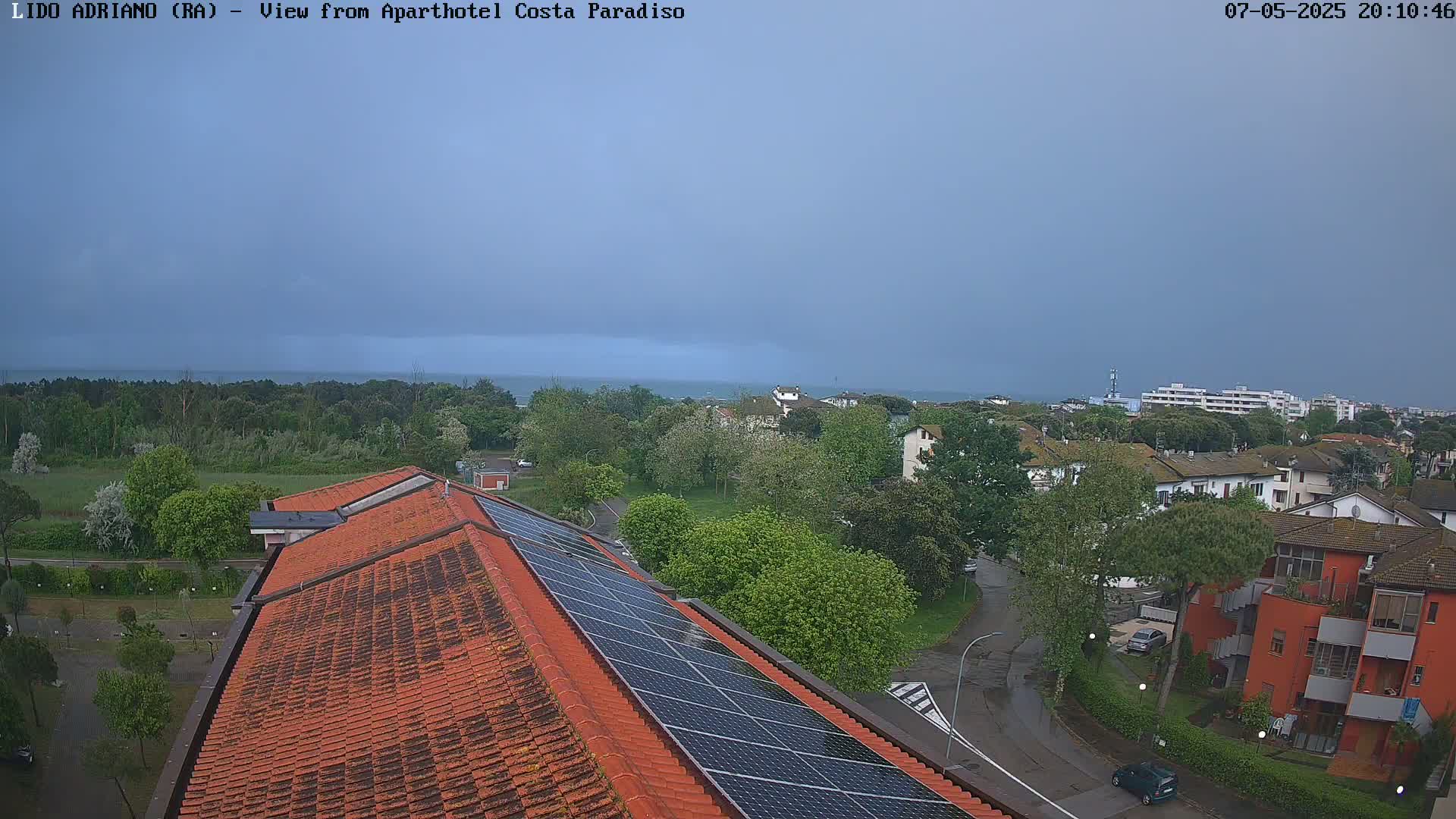 A view of a coastal town under an overcast sky shows a red tiled roof with solar panels, a wet street, and lush green trees, with a calm sea visible in the distance.