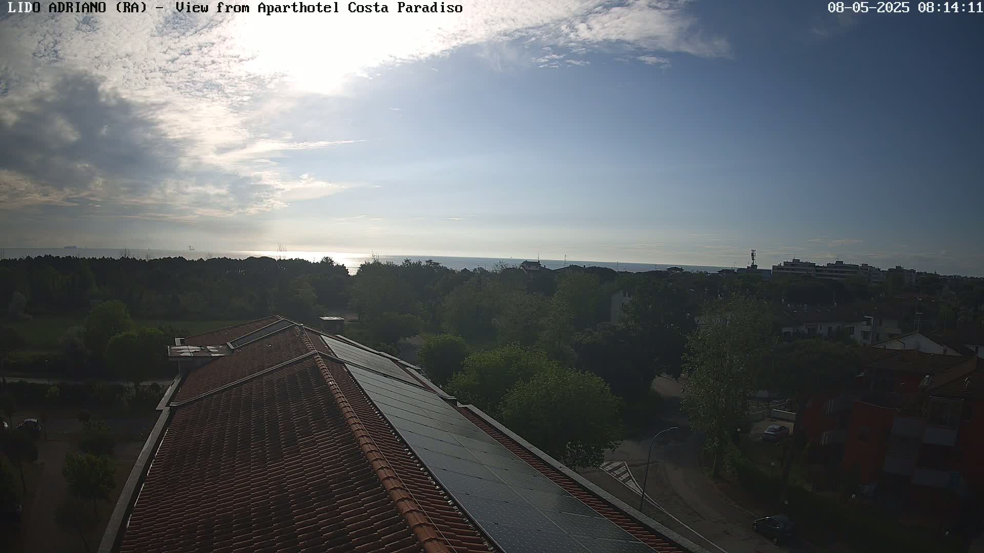 A partly cloudy sky overlooks a tiled roof with solar panels, green trees, and a calm sea in the distance.