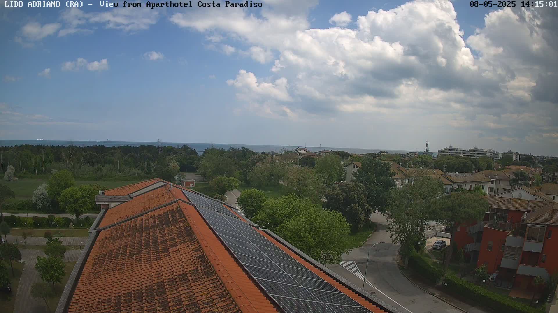 A partly cloudy sky overlooks a coastal town with red-tiled rooftops and solar panels, a calm sea in the distance.