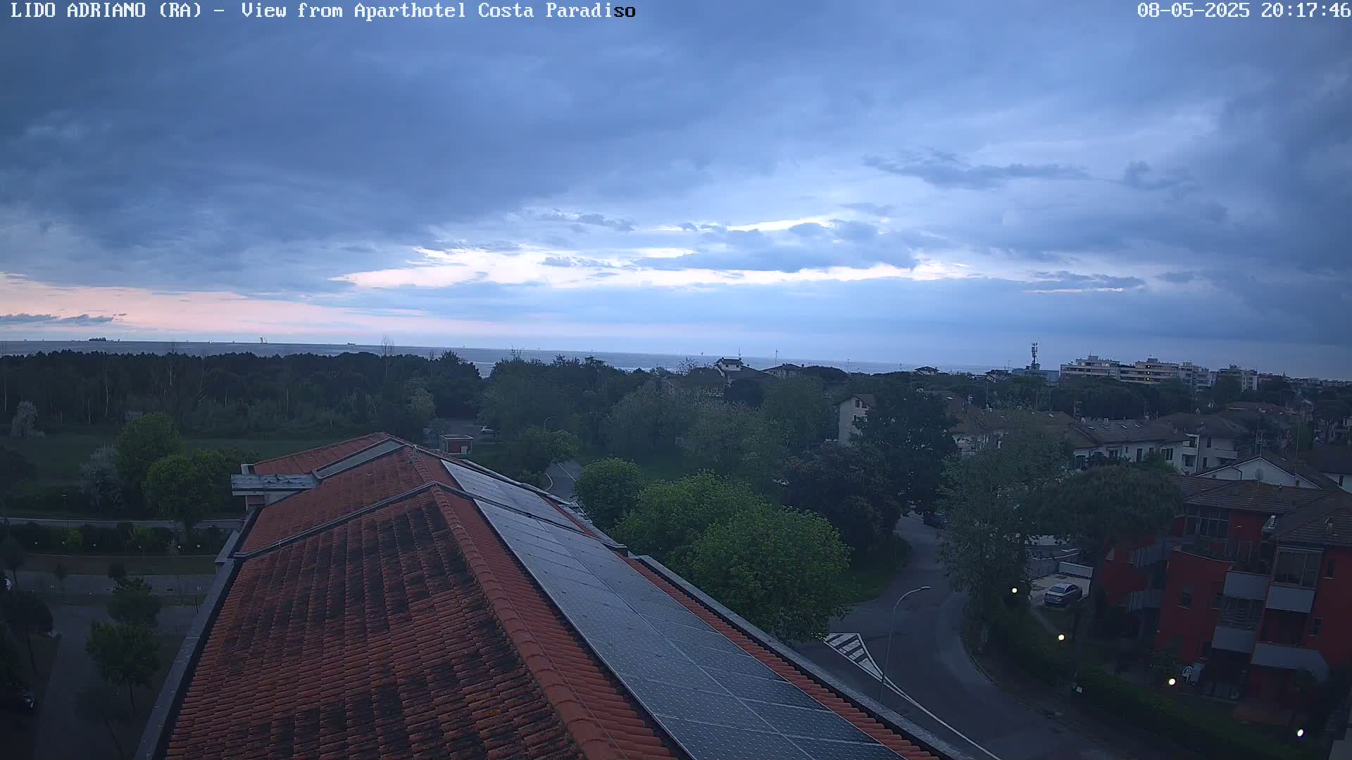 A view from a rooftop shows a partly cloudy evening sky over a residential area and a calm sea in the distance.