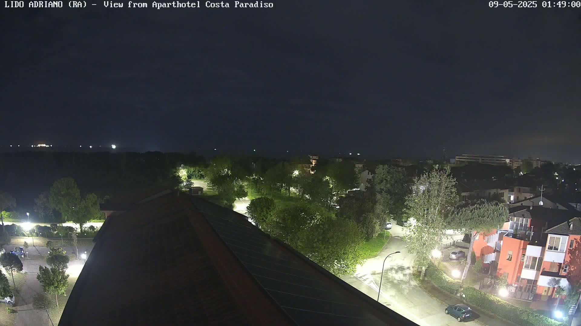 A nighttime view from a rooftop shows a residential area with streetlights illuminating trees, buildings, and roads under a dark sky.