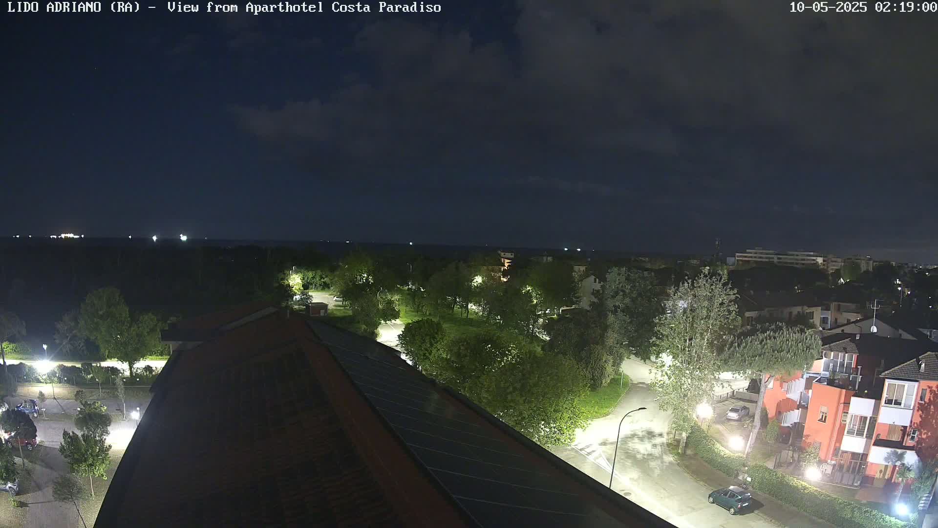 A nighttime, overhead view of a residential area near a body of water under a partly cloudy sky shows a dark ocean, illuminated trees, streetlights, and buildings.