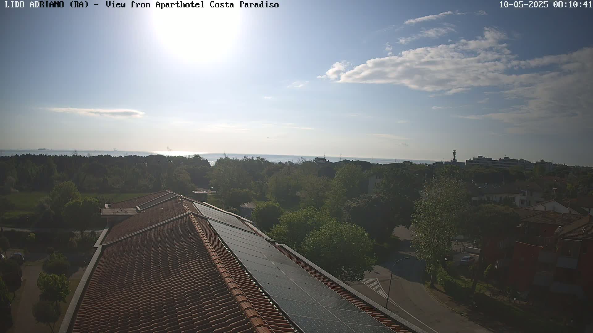 A partly cloudy sky overlooks a seaside town with green trees, buildings, and a red tiled roof with solar panels.