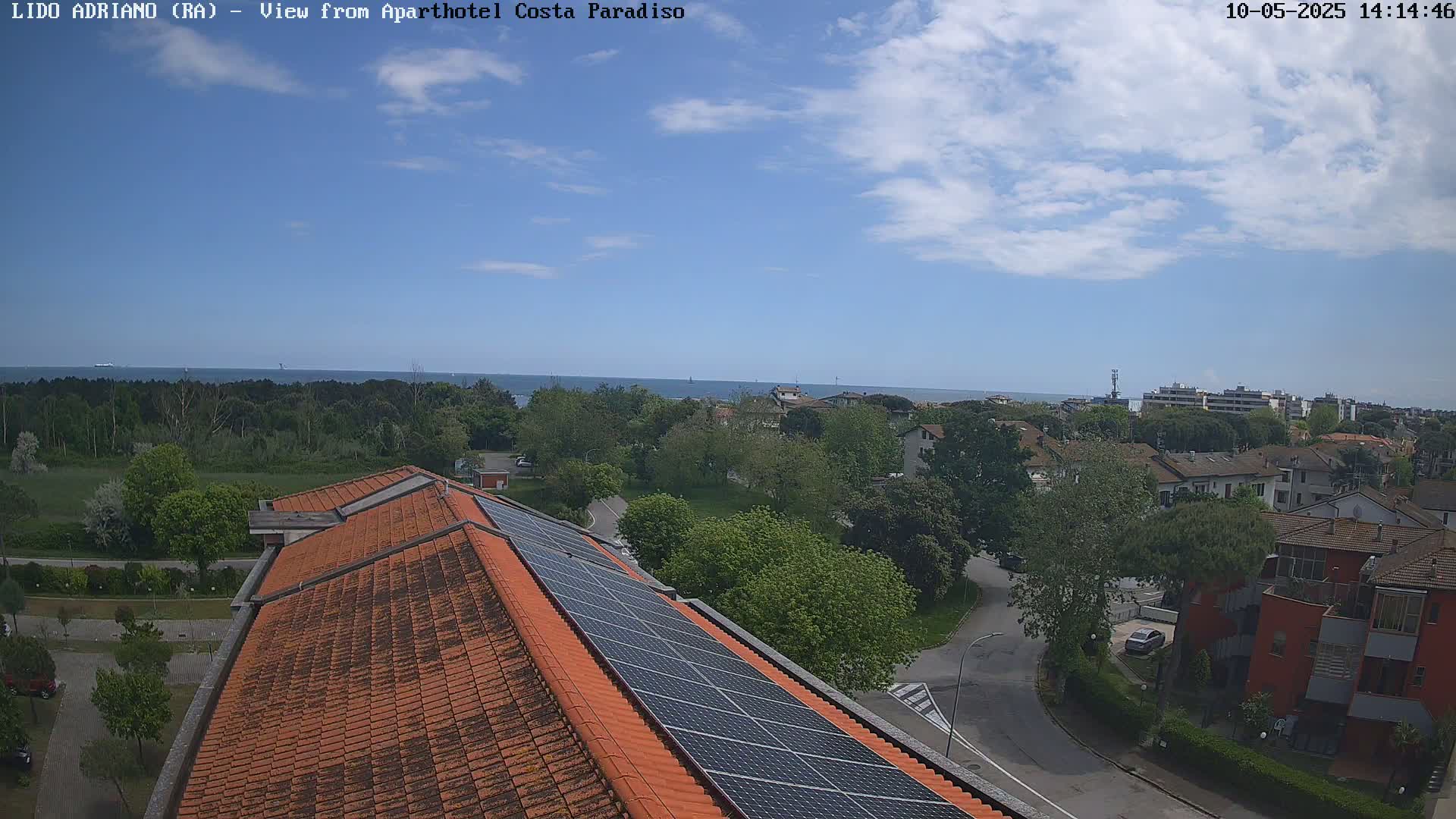 An overhead view shows a partly cloudy sky over a coastal town with green trees, residential buildings, and a building with solar panels on its orange tile roof; a calm sea is visible in the distance.