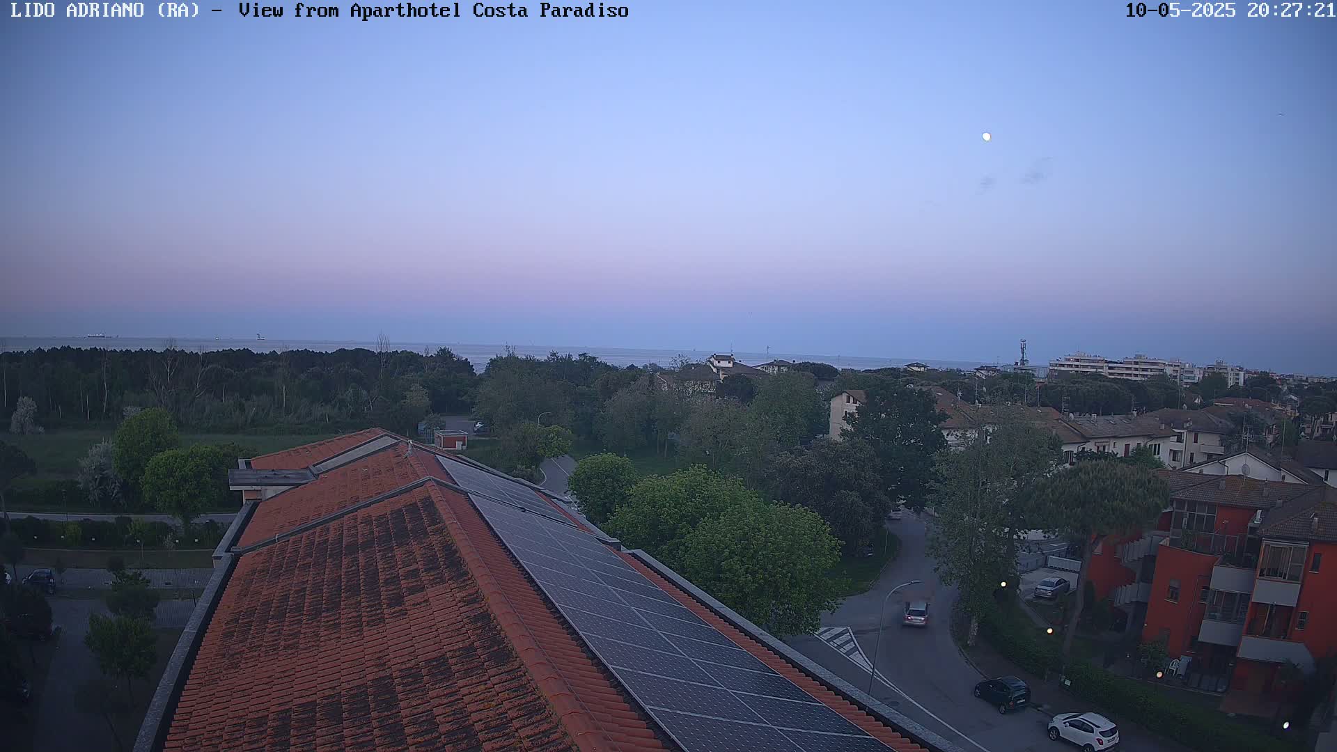 A twilight view from a rooftop with solar panels shows a calm sea, a line of trees, and a residential area under a nearly full moon.