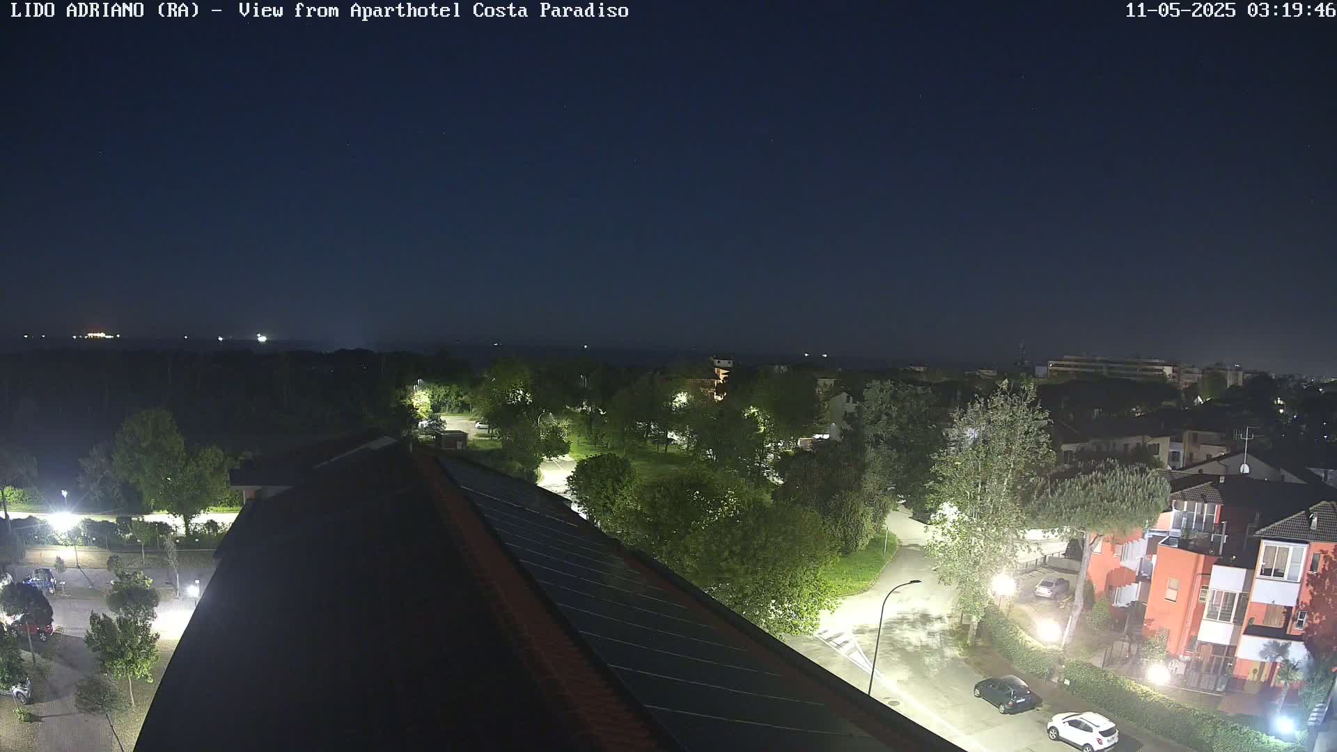 A nighttime view from a rooftop shows a residential area with streets, trees, and buildings illuminated by streetlights, under a clear, dark sky.
