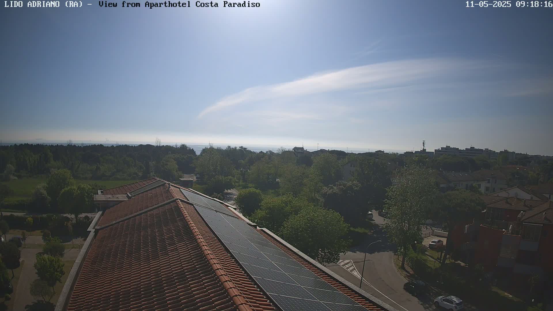 A mostly sunny day overlooks a tiled roof with solar panels, green trees, residential buildings, and a calm body of water in the distance.