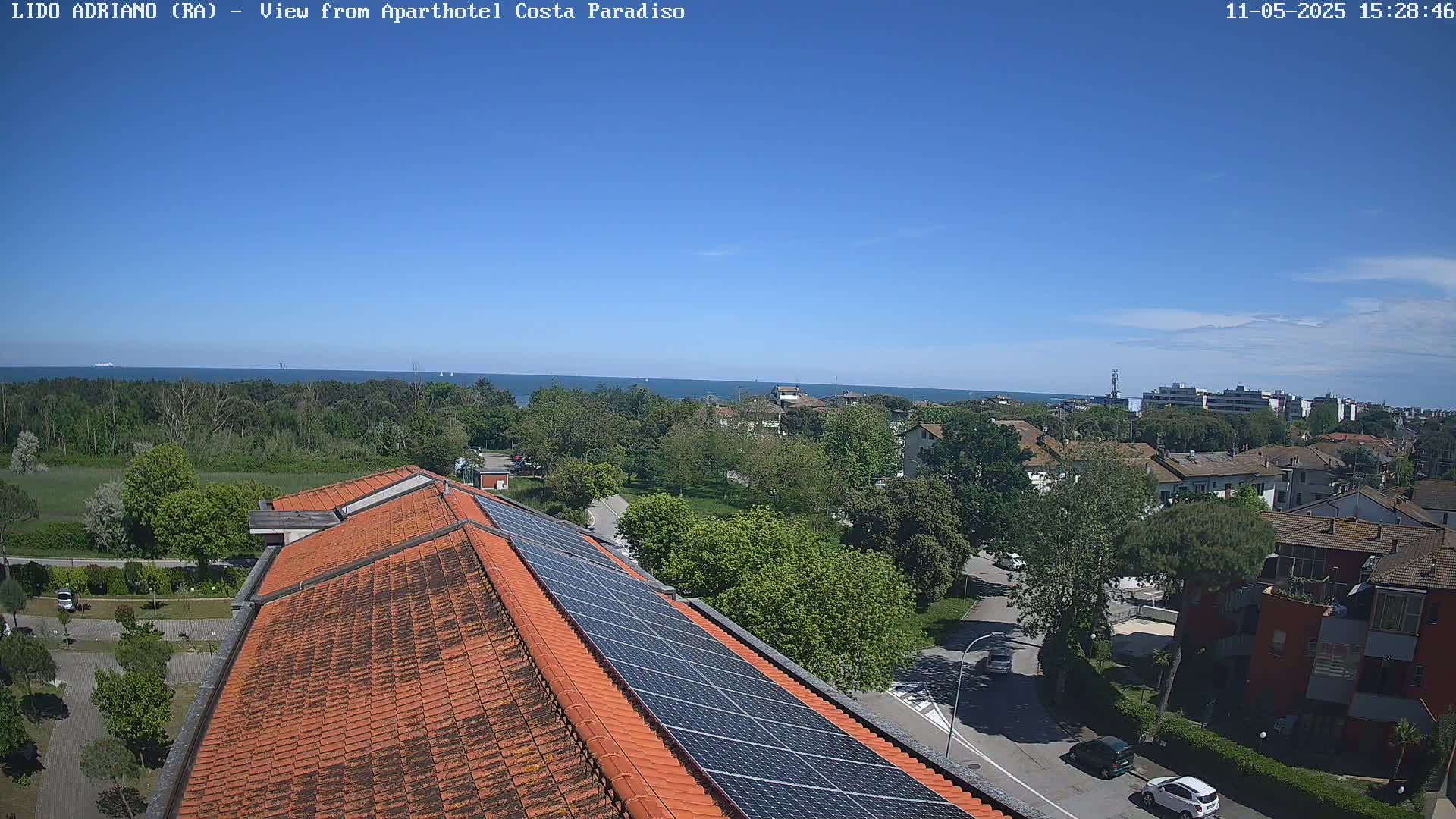 A clear, sunny day reveals a coastal town scene viewed from a rooftop with solar panels, overlooking a calm sea and lush green trees.