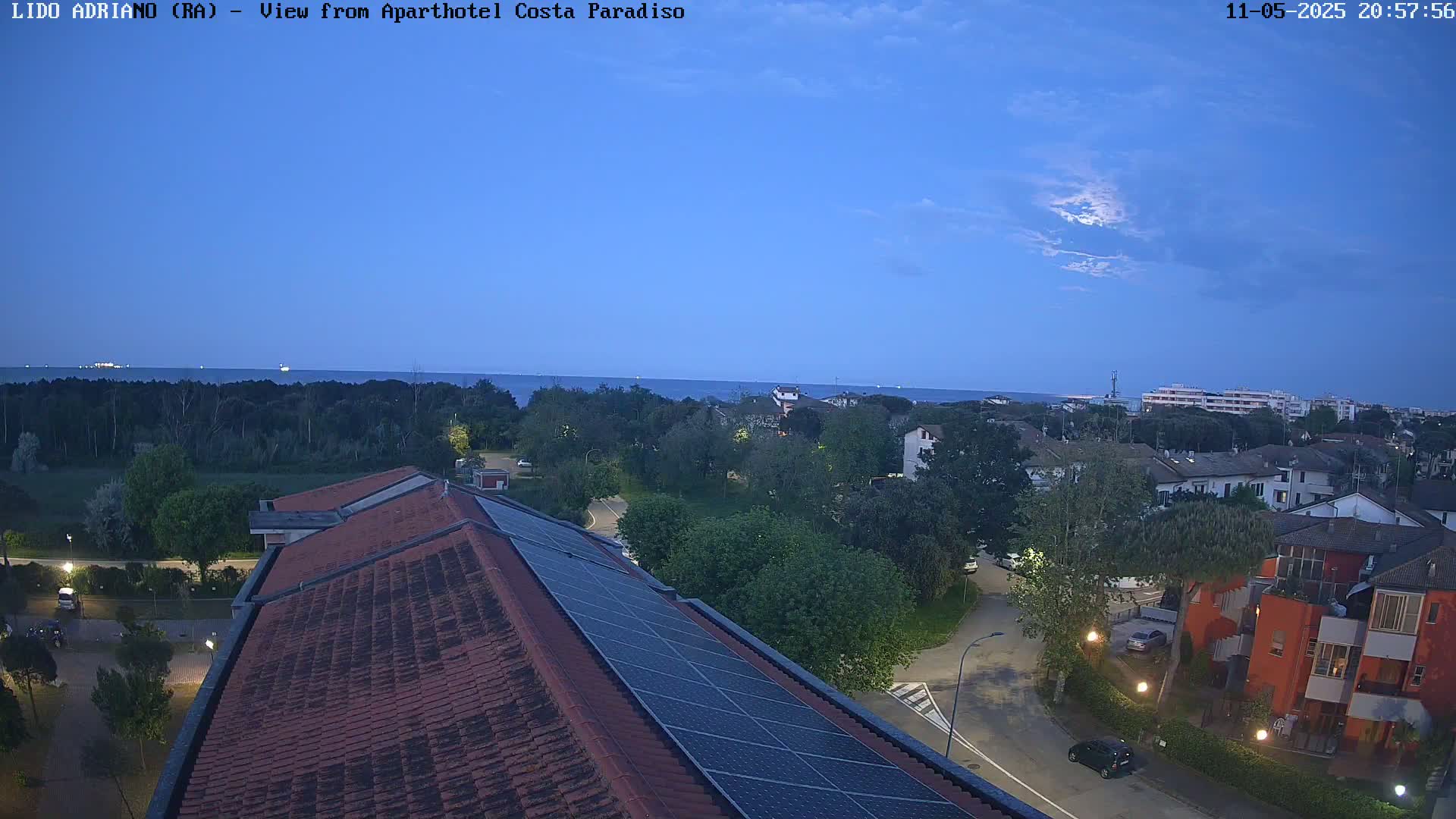 A rooftop with solar panels overlooks a calm twilight sea, a strip of trees, and residential buildings under a partly cloudy sky.